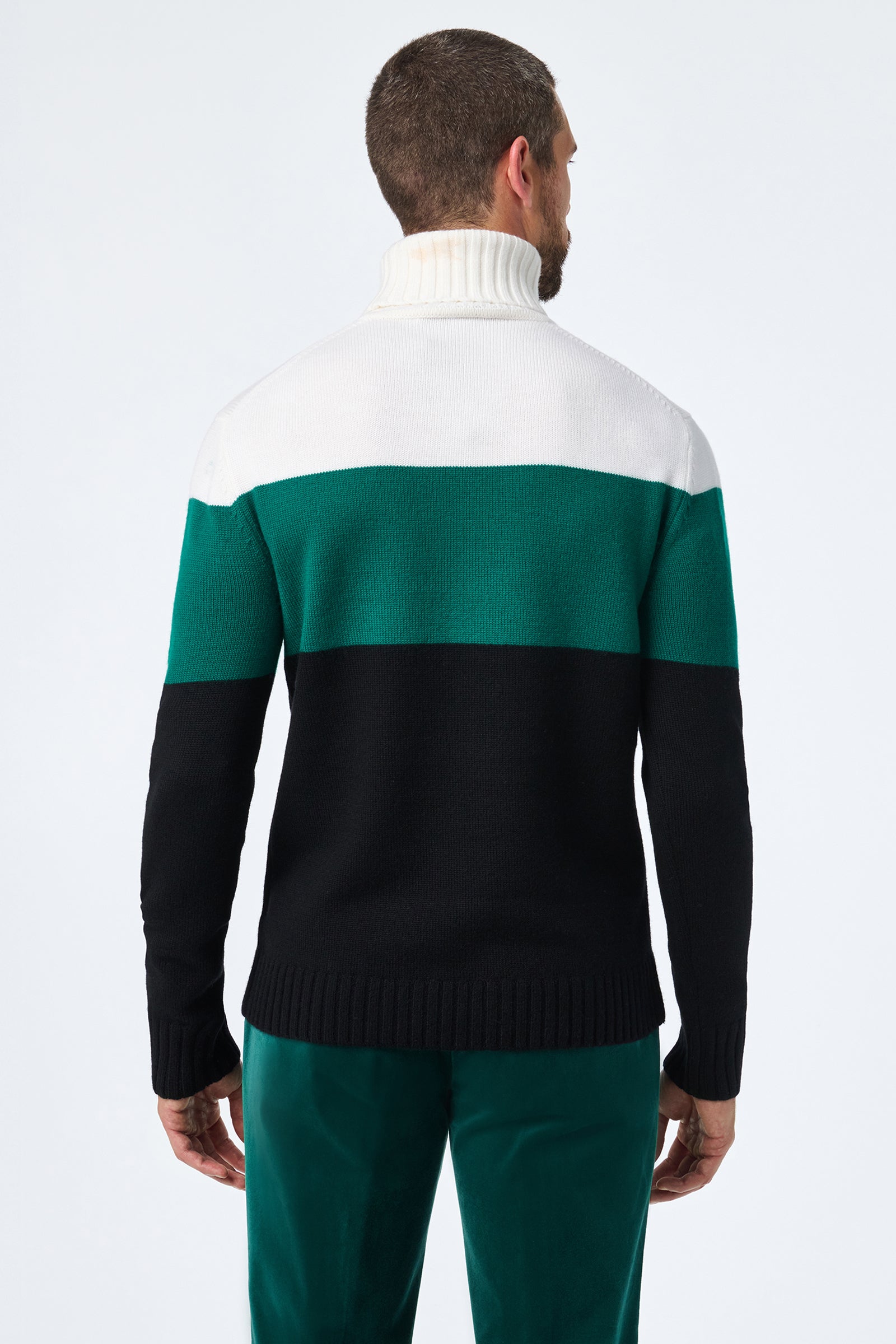 A man faces away wearing the Harrelson T-Neck, a color-blocked turtleneck sweater with white, green, and black horizontal sections, paired with green pants against a plain light background.