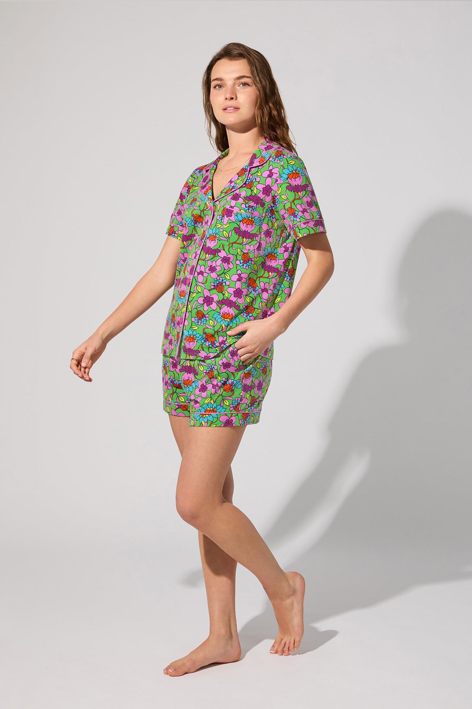 A woman stands barefoot against a plain background, wearing the Shorty PJ Set - Garden Floral—a matching short-sleeve button-up top and shorts with a green, pink, and purple floral pattern. She has one hand in her pocket and looks forward.