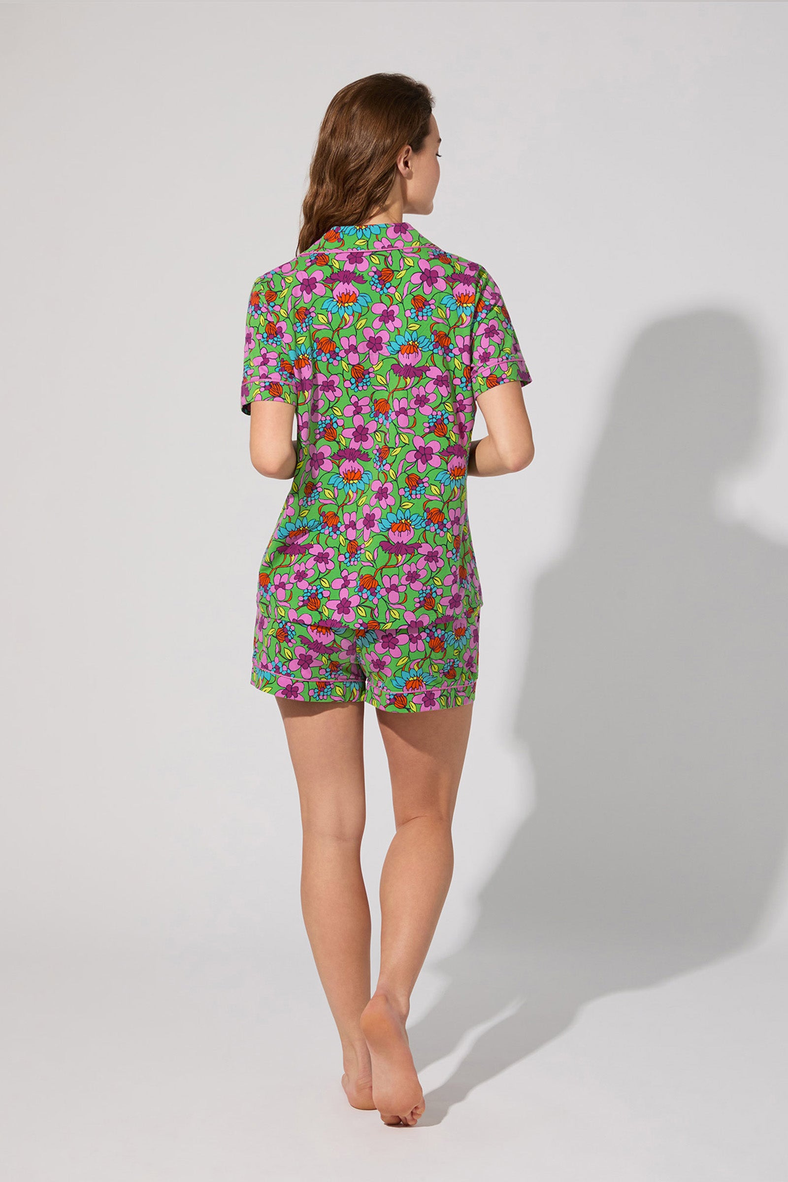 A woman stands barefoot facing away, wearing the Shorty PJ Set - Garden Floral, a matching short-sleeve shirt and shorts with a vibrant floral pattern on a plain light background.