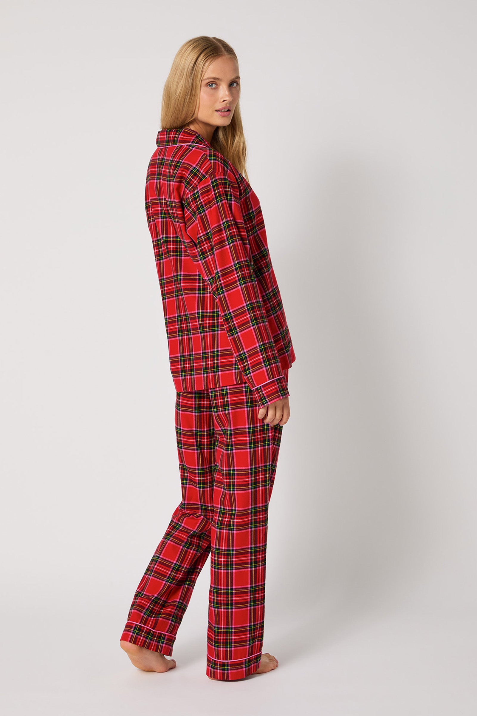 A woman with long blonde hair stands barefoot, wearing the Christmas Plaid L/S Classic PJ, facing sideways and looking over her shoulder against a plain light background.