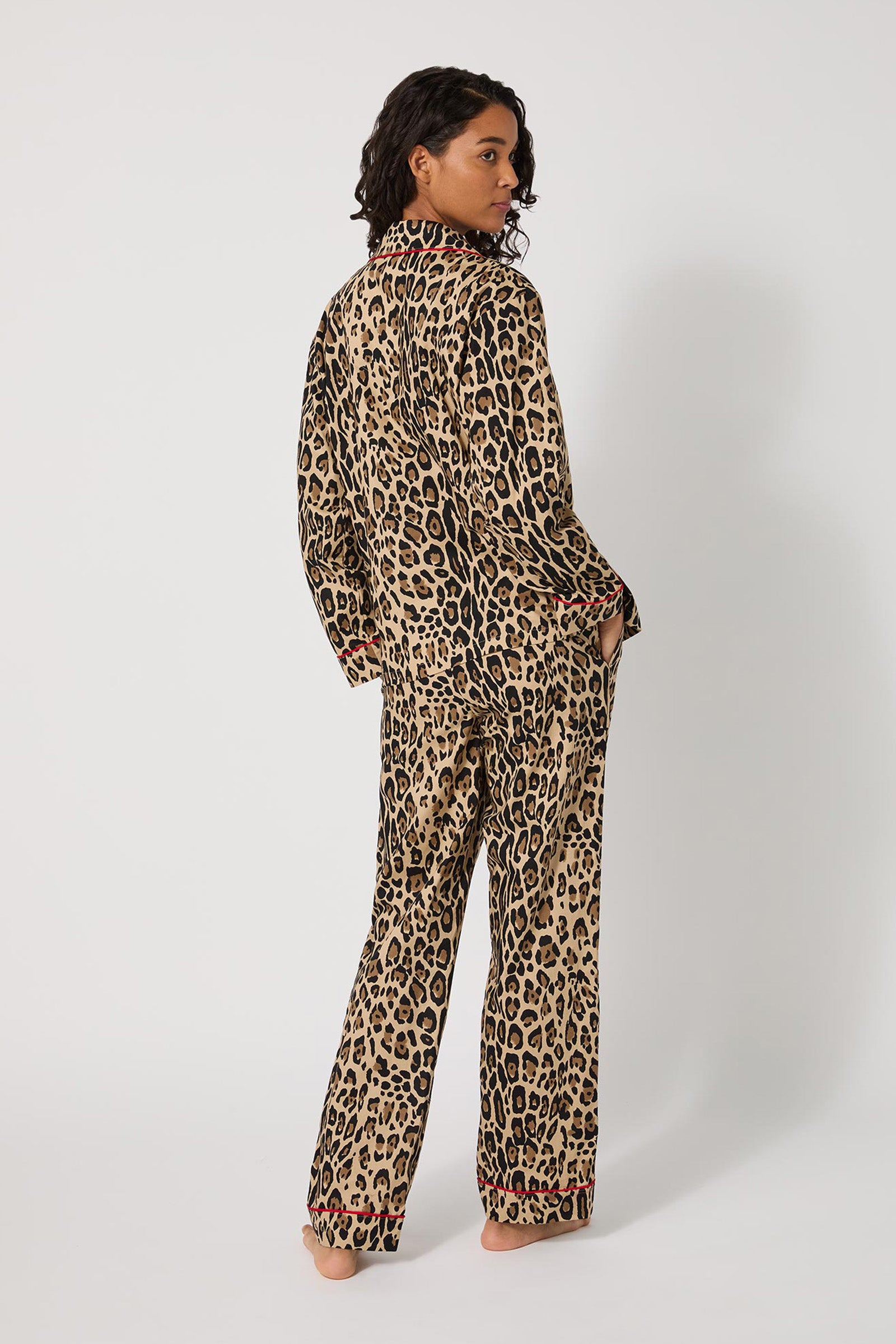 A person with curly dark hair stands barefoot, modeling the Leopard Stripe L/S Classic Pj set in tan and black leopard print, glancing over their shoulder against a plain white background.