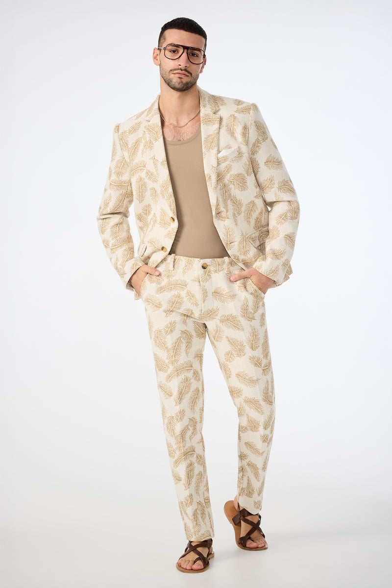 A man poses confidently against a plain white background, hands in pockets, wearing the Alex Slim Trouser in Desert Fronds Jacquard paired with a tan shirt, brown sandals, and large glasses.