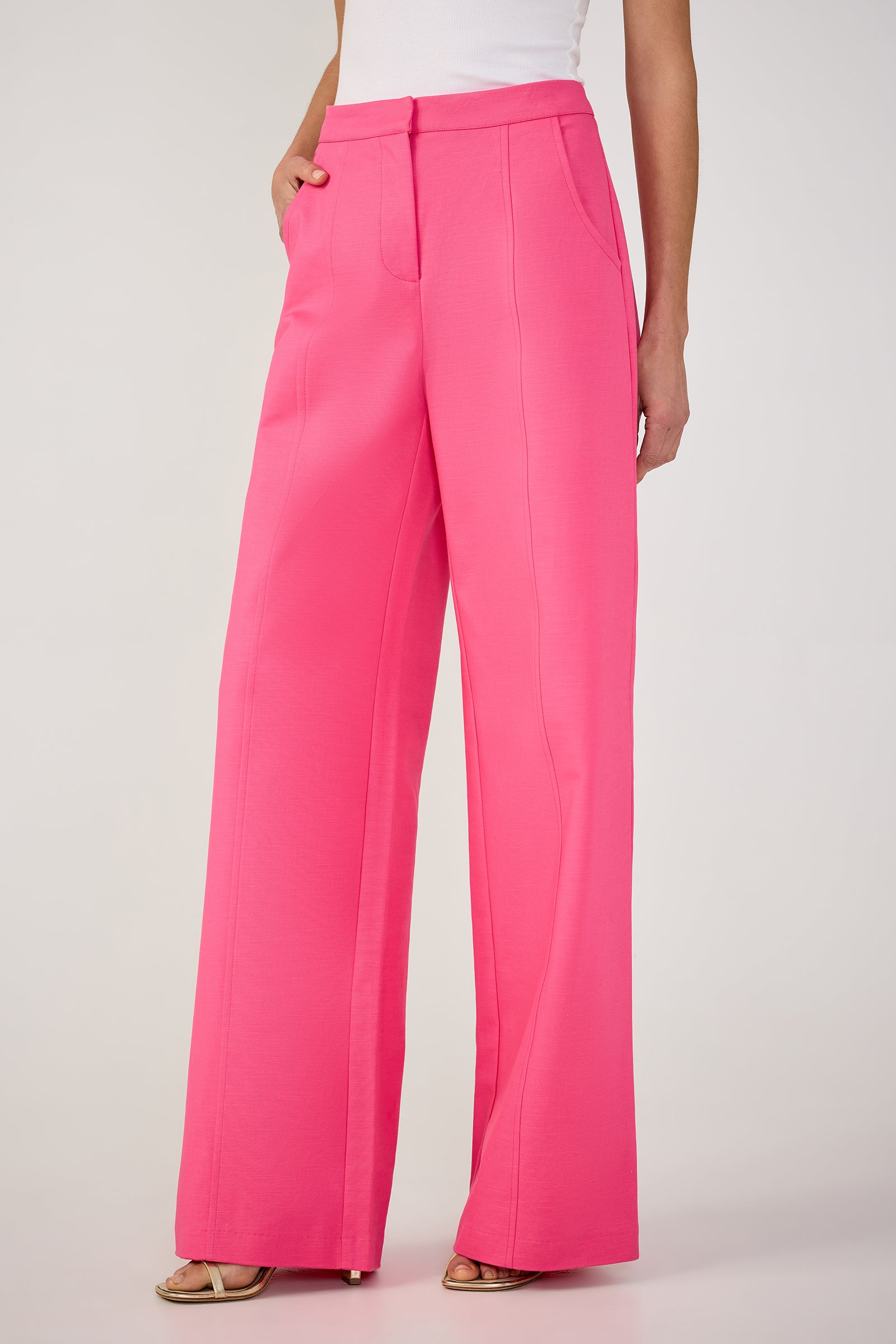 A person wears the Half Court Wide-Leg Pant - Solid Suiting in bright pink, paired with a white sleeveless top and open-toed heels, standing with one hand in a pocket against a plain light background.
