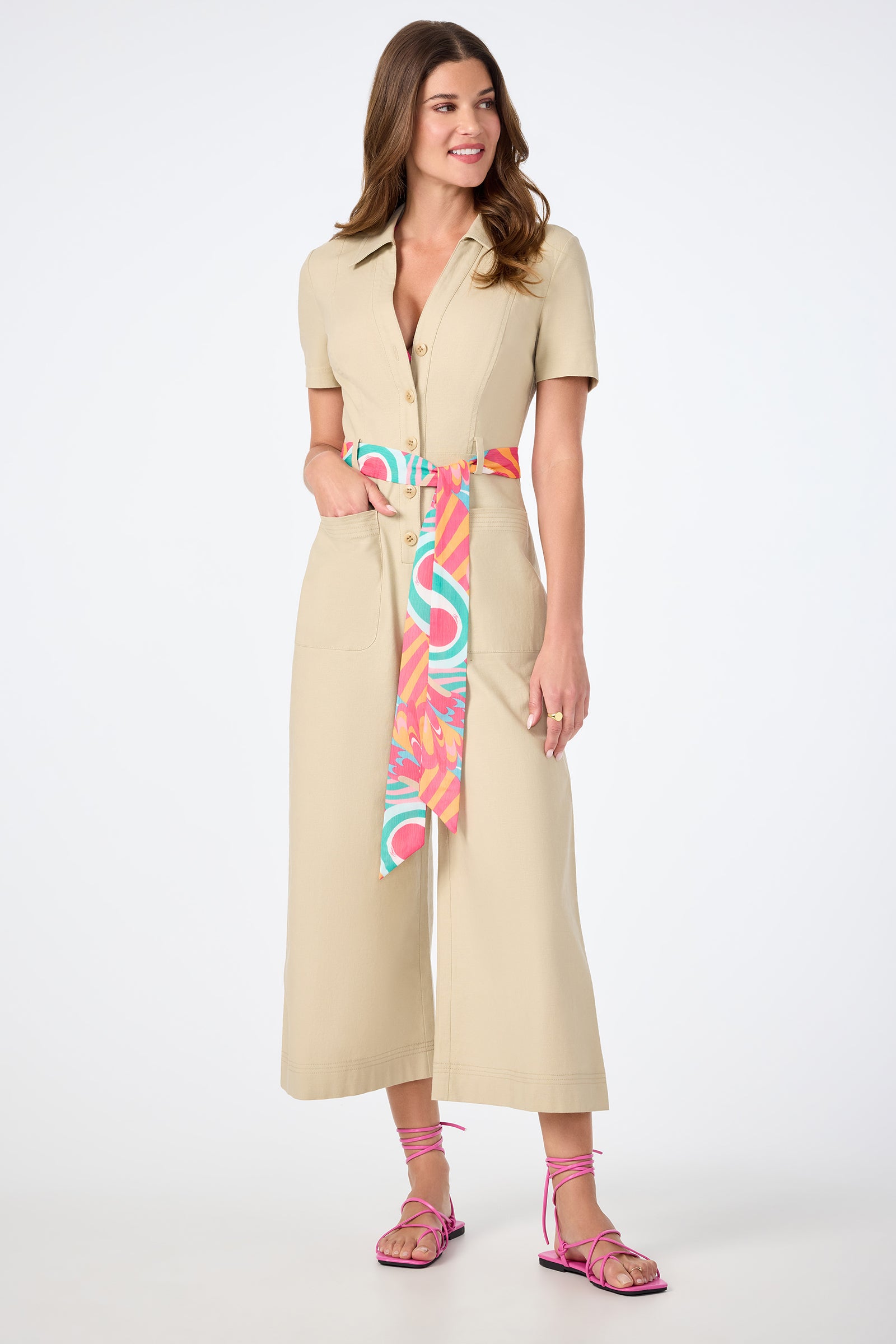 A woman smiles against a plain white background, wearing the JAdore V-Neck Jumpsuit in beige with short sleeves, wide legs, a colorful sash belt, and pink strappy sandals.