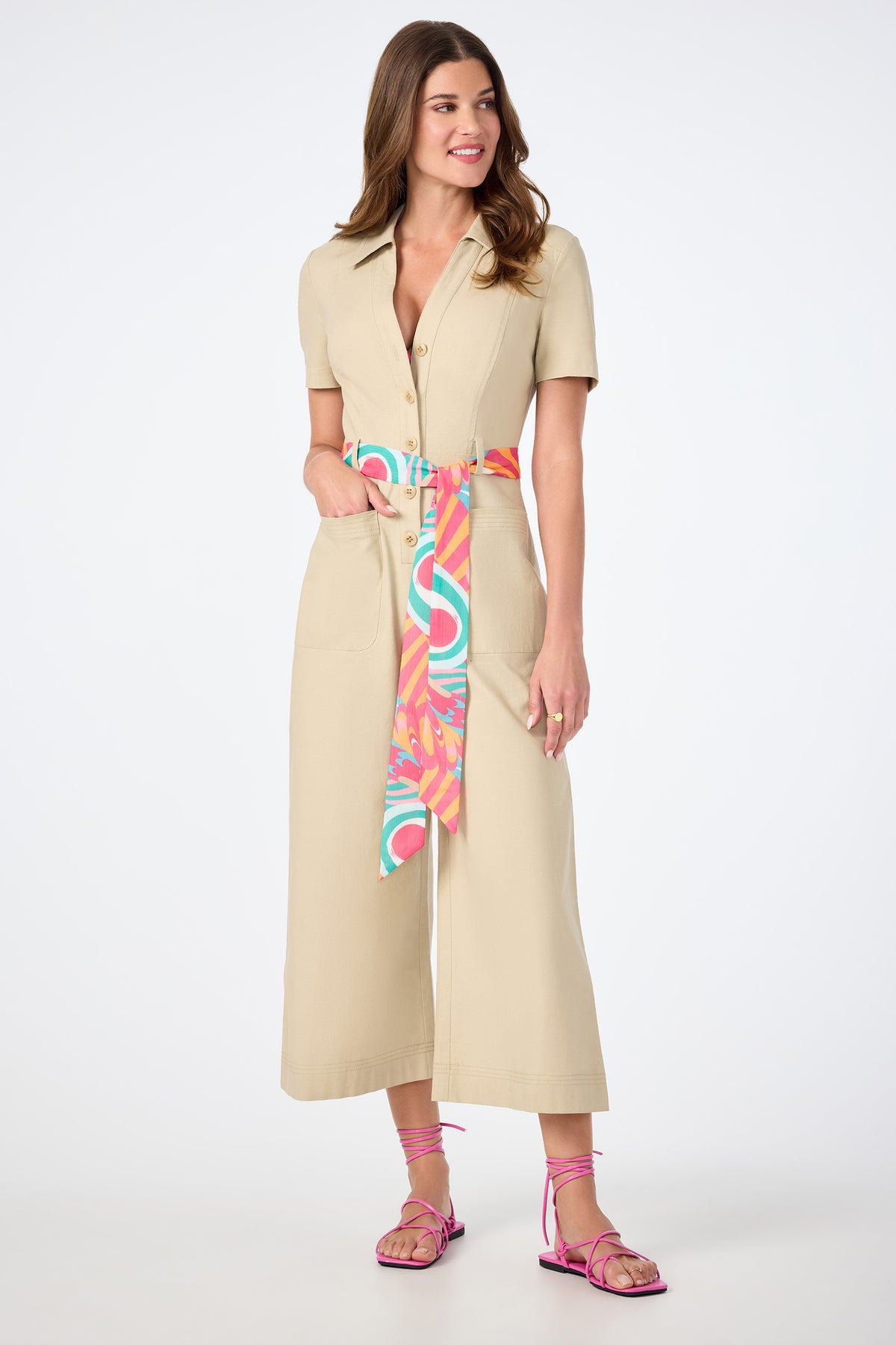A woman smiles against a plain white background, wearing the JAdore V-Neck Jumpsuit in beige with short sleeves, wide legs, a colorful sash belt, and pink strappy sandals.