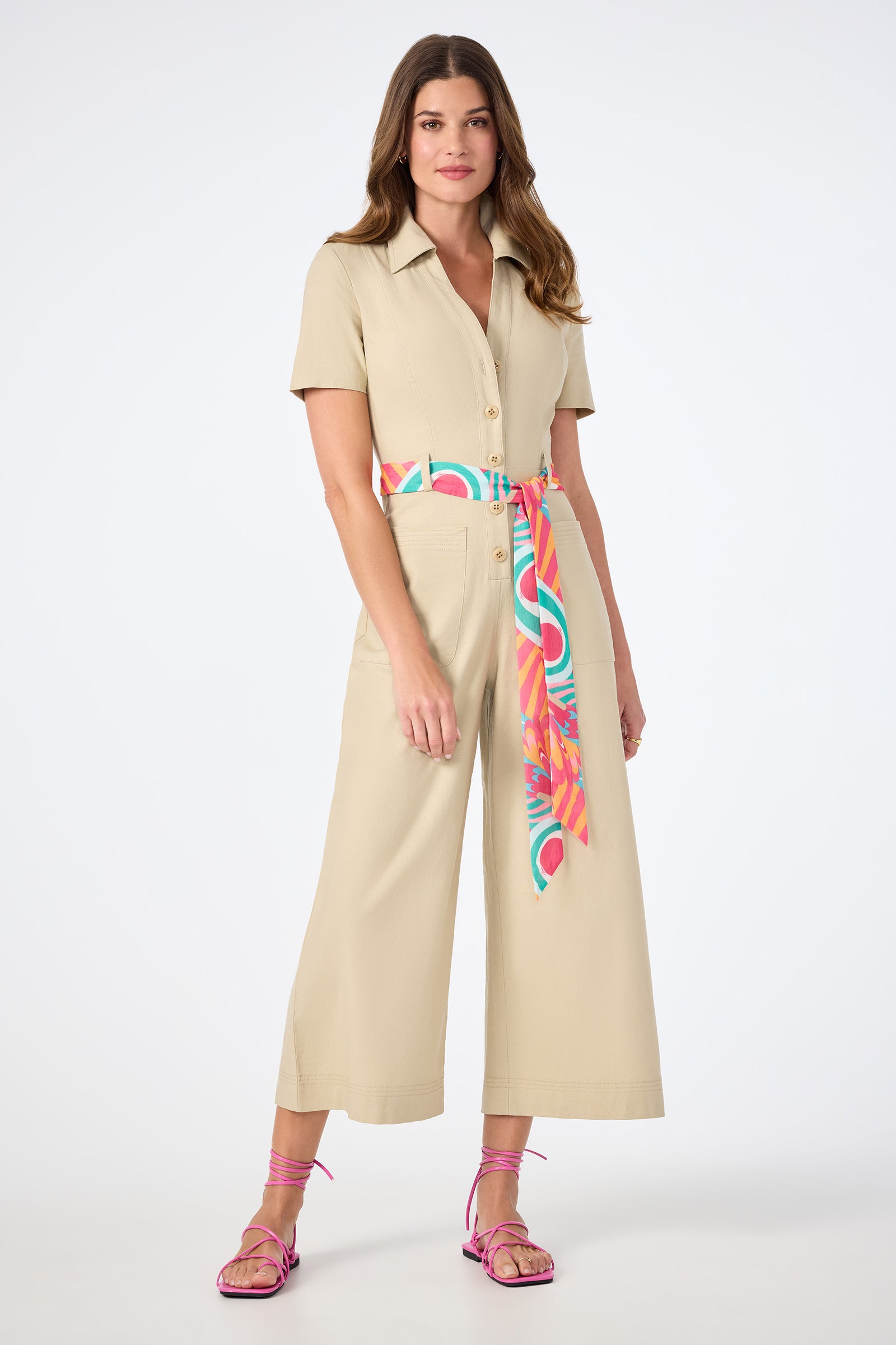 A woman with long, wavy hair and a neutral expression poses against a white background, wearing the JAdore V-Neck Jumpsuit in Biarritz Beige, styled with a colorful sash belt and pink strappy sandals.