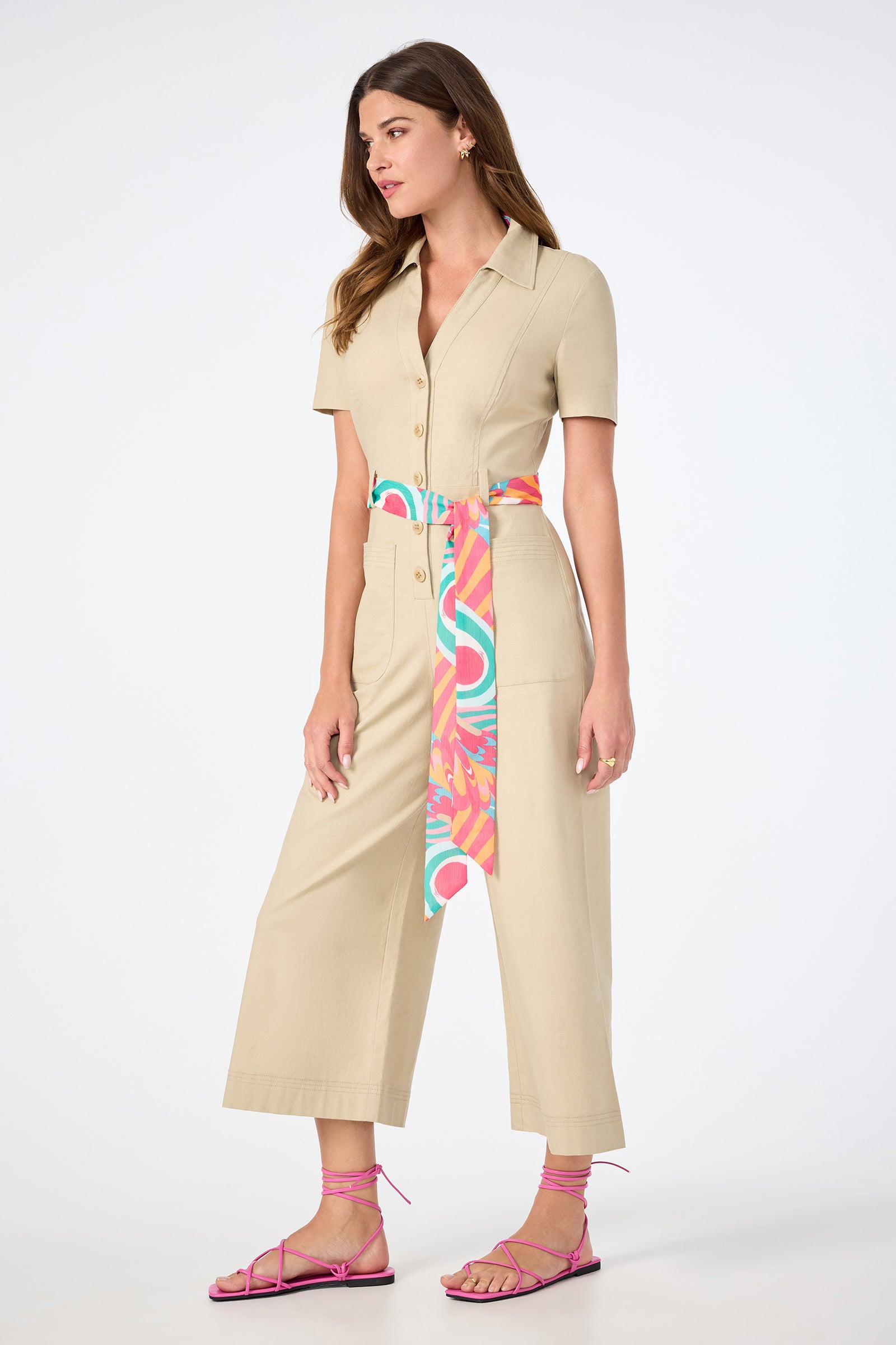 A woman in the JAdore V-Neck Jumpsuit, featuring beige short sleeves and wide legs, styled with a colorful scarf belt and pink strappy sandals, stands against a plain white background.