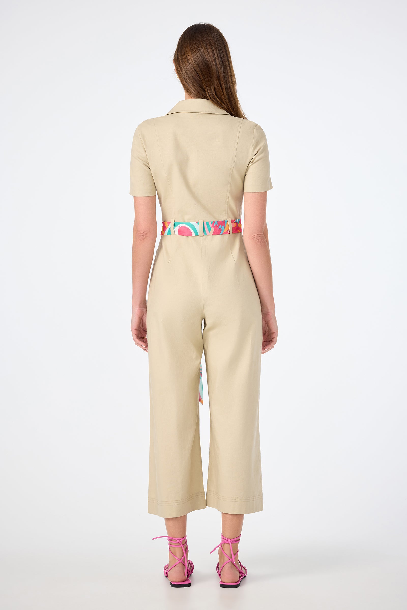 A woman faces away, wearing the JAdore V-Neck Jumpsuit in beige with short sleeves and wide legs, styled with a colorful print belt and pink strappy sandals. Her straight brown hair is down against a plain white background.