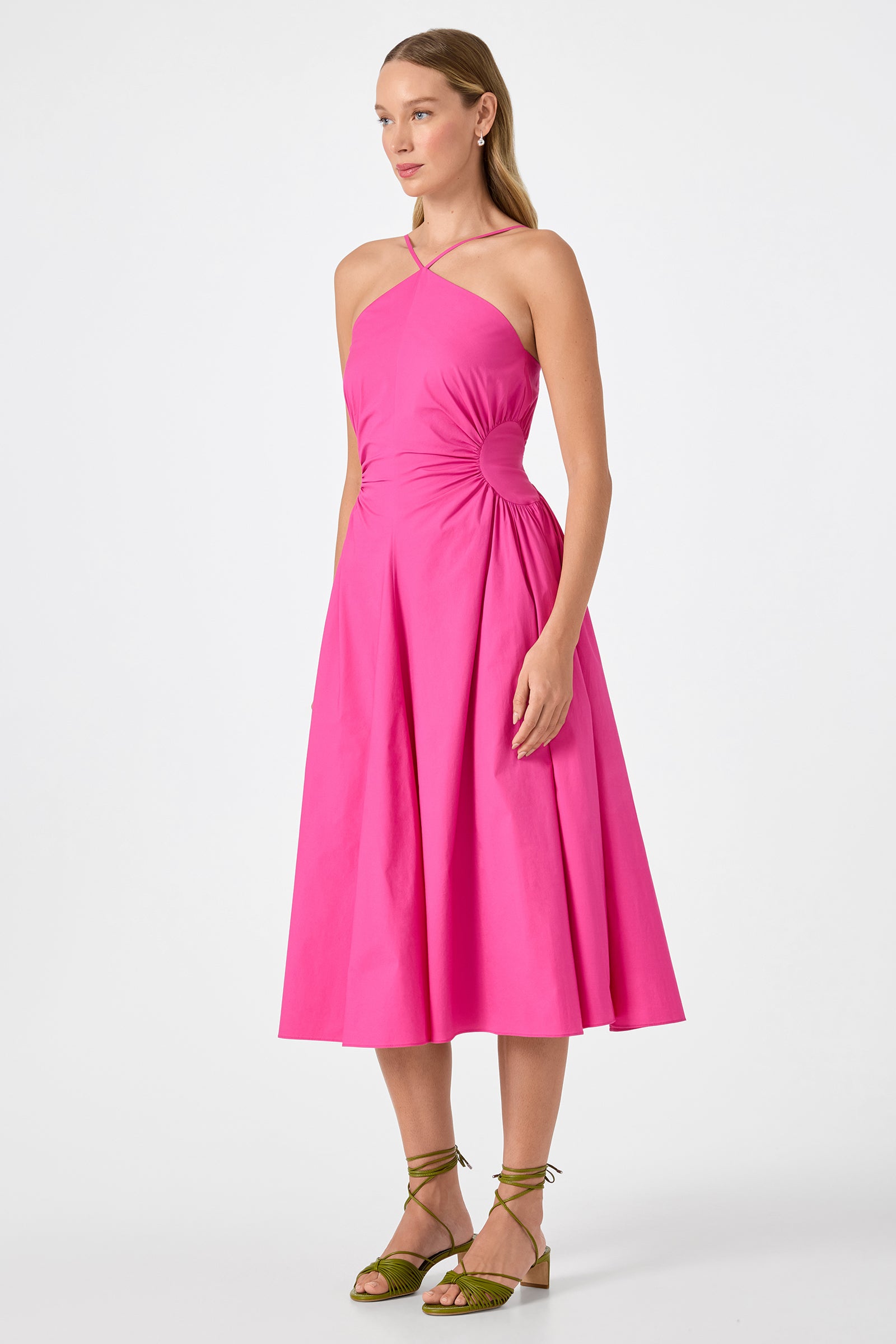 A woman stands against a plain white background wearing the Kata Shirred Waist Midi Dress, a bright pink, sleeveless midi with a crisscross halter neckline and ruched sides, paired with olive green strappy sandals.