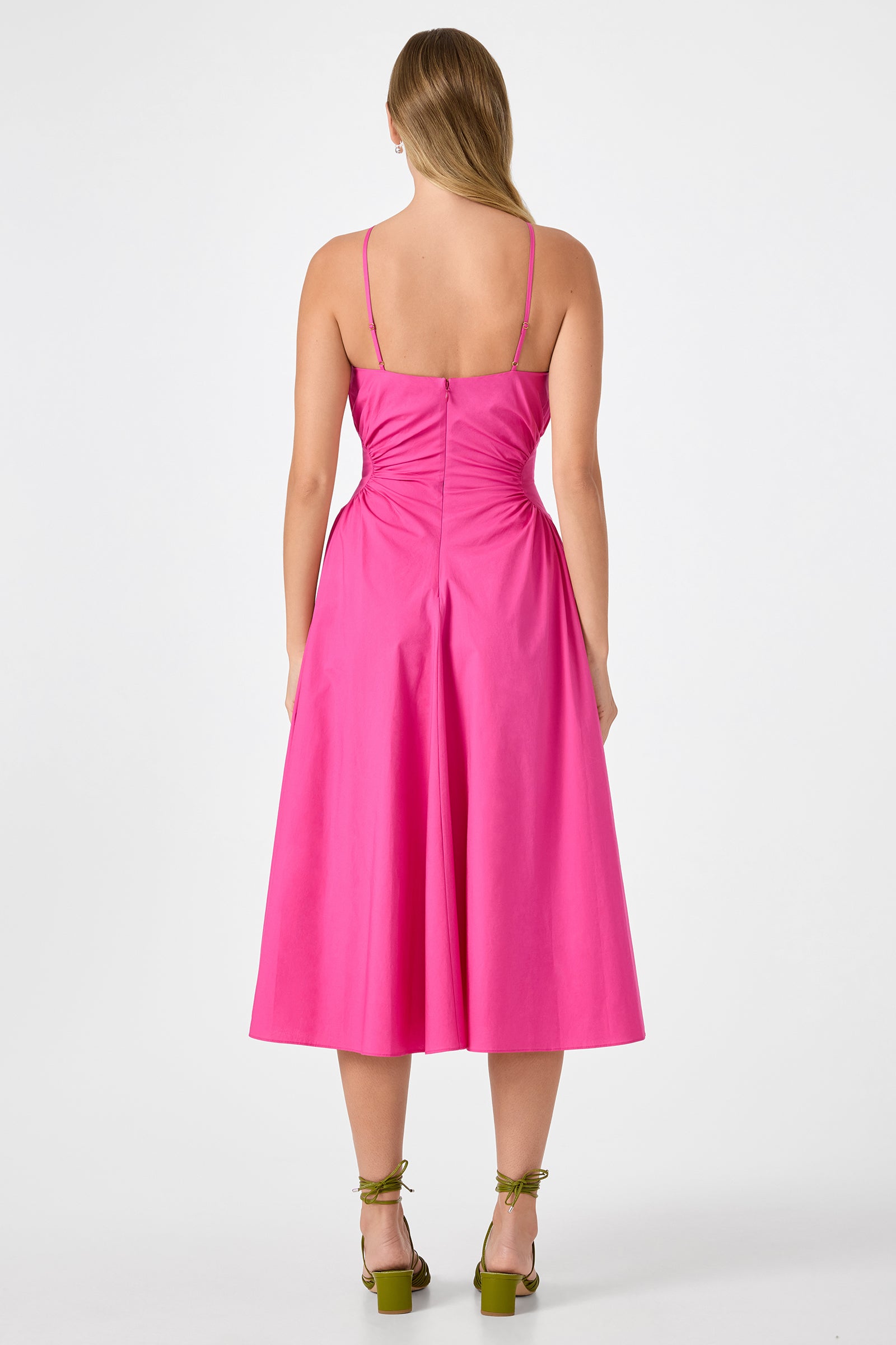 A woman with light brown hair faces away, wearing the Kata Shirred Waist Midi Dress in bright pink. The sleeveless, mid-length dress features thin straps and back ruching. She pairs it with green open-toe ankle-strap heels.