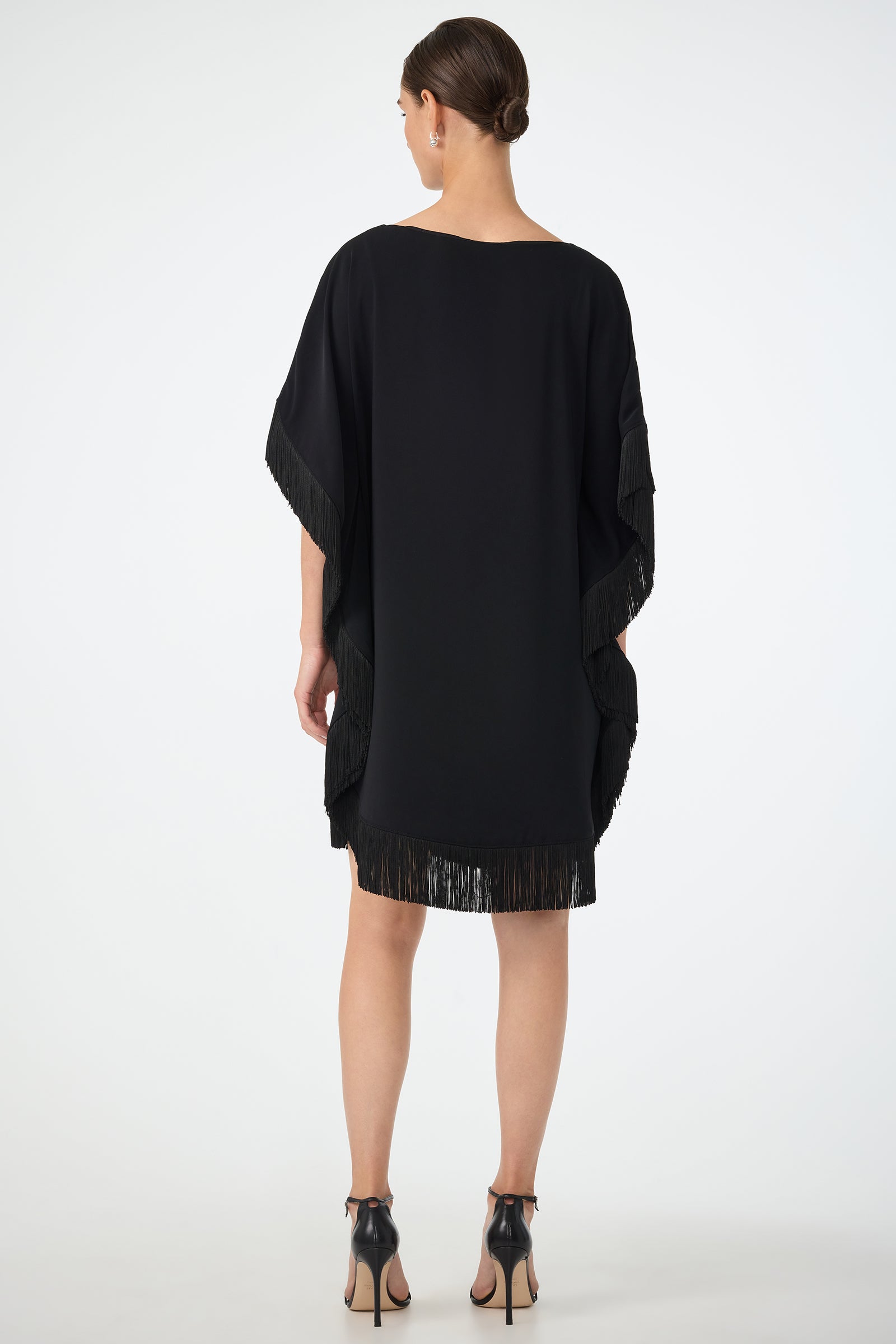 A woman stands facing away, wearing the Mini Neena Fringe Caftan Dress—a loose black dress with fringe at the sides and hem—paired with black high-heeled sandals. Her hair is styled in a neat low bun.