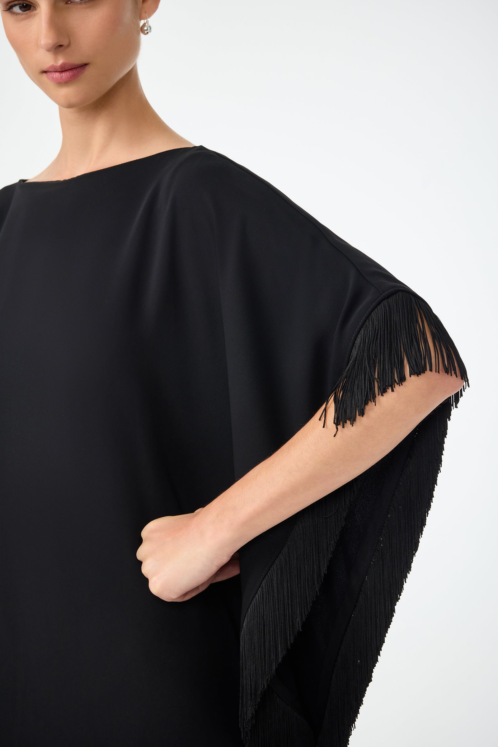 A woman models the Mini Neena Fringe Caftan Dress in black, standing against a plain background with her hand on her hip. The cropped image partially shows her face and highlights the dresss fringed sleeves.