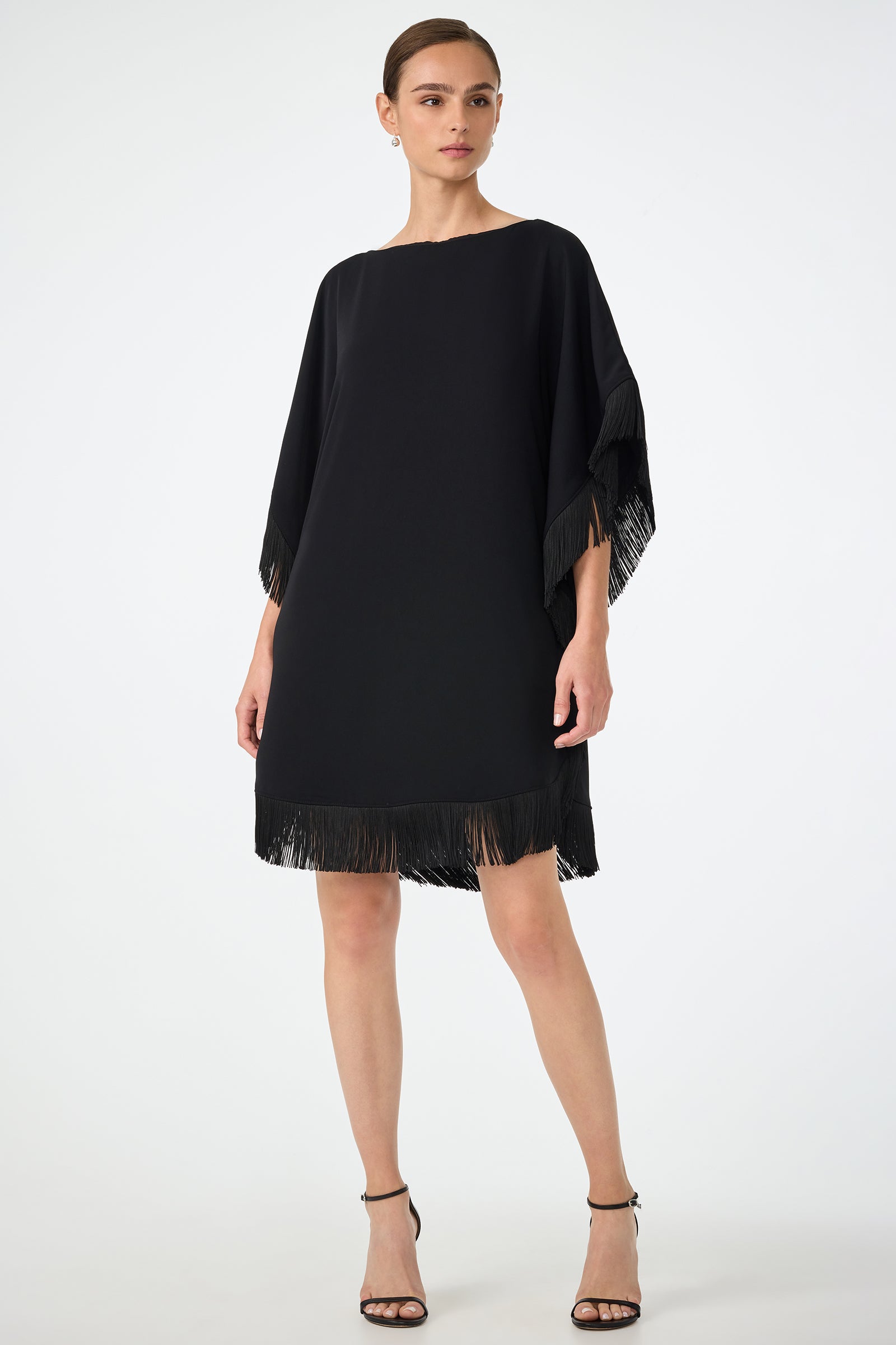 A woman models the Mini Neena Fringe Caftan Dress in black, featuring loose fit and fringe on the sleeves and hem. She pairs it with black high-heeled sandals and stands confidently against a plain white background.