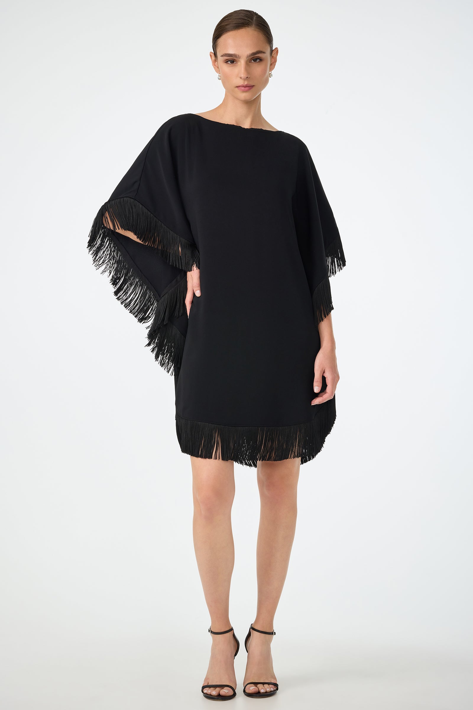 A woman wears the Mini Neena Fringe Caftan Dress—loose-fitting, knee-length, black with fringe on sleeves and hem—paired with black high-heeled sandals against a plain light background.