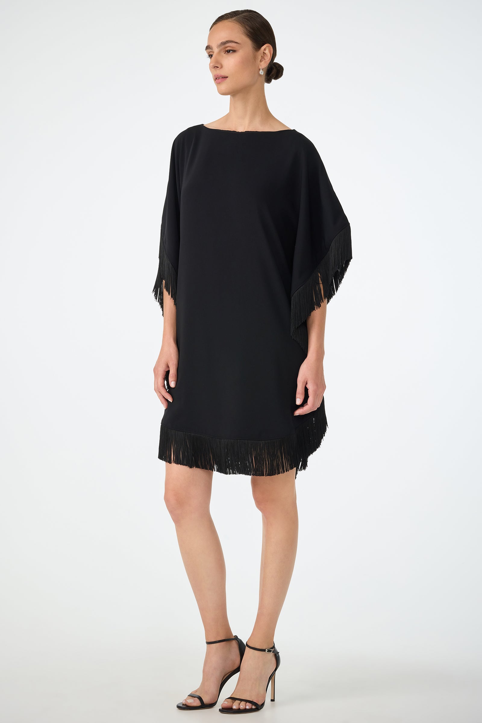 A woman wearing the Mini Neena Fringe Caftan Dress, a loose-fitting black dress with fringe details on the sleeves and hem, paired with black high-heeled sandals, stands against a plain light background.