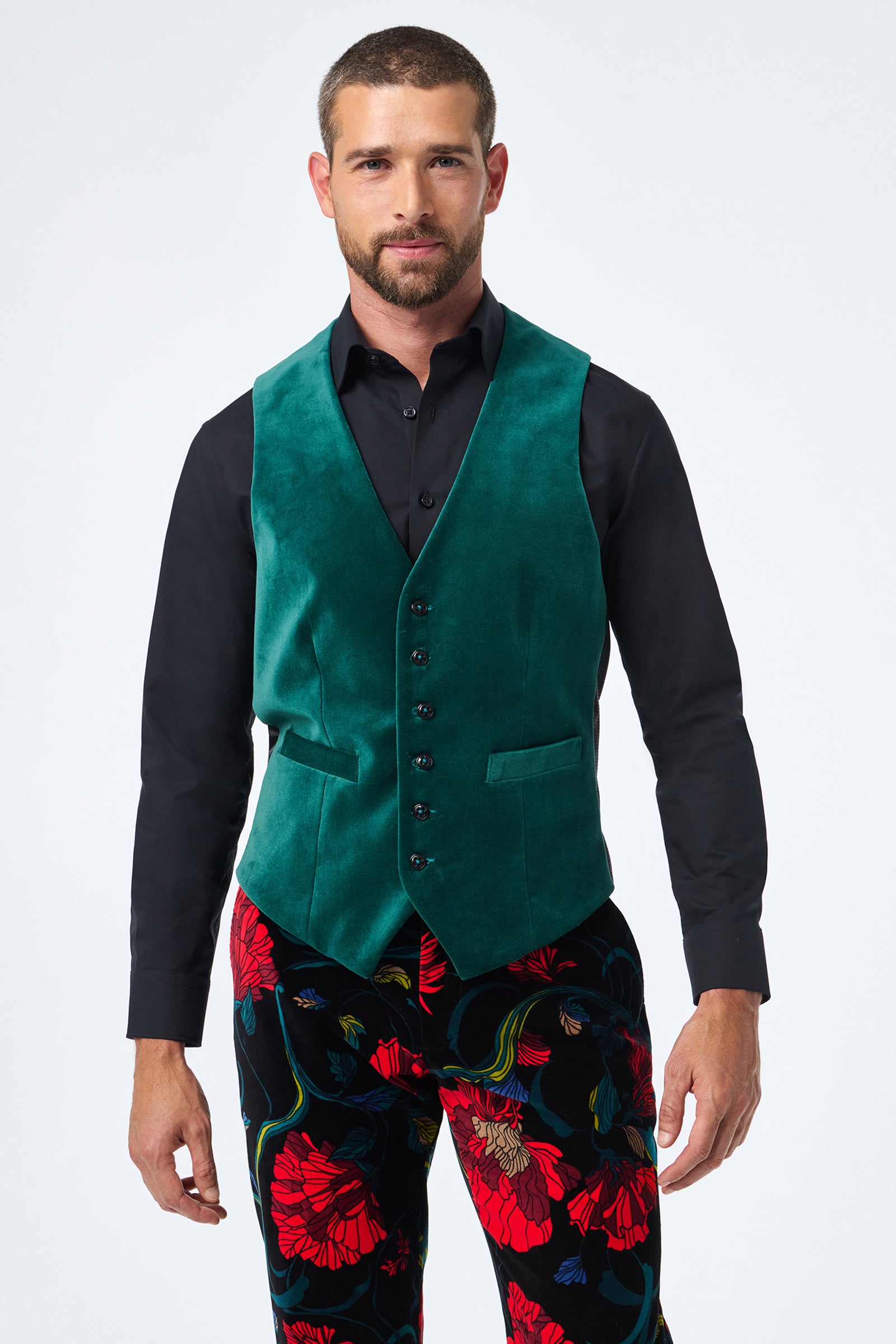 A man wearing the Irving Vest in green velvet over a black button-up shirt and colorful floral-patterned pants stands against a plain light background.