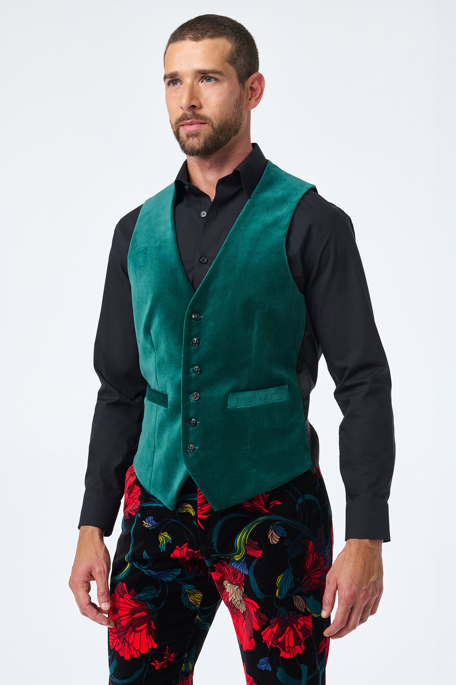 A man with a short beard wears the Irving Vest, a green velvet piece, over a black button-up shirt and black pants with a red and blue floral pattern, standing against a plain light background.