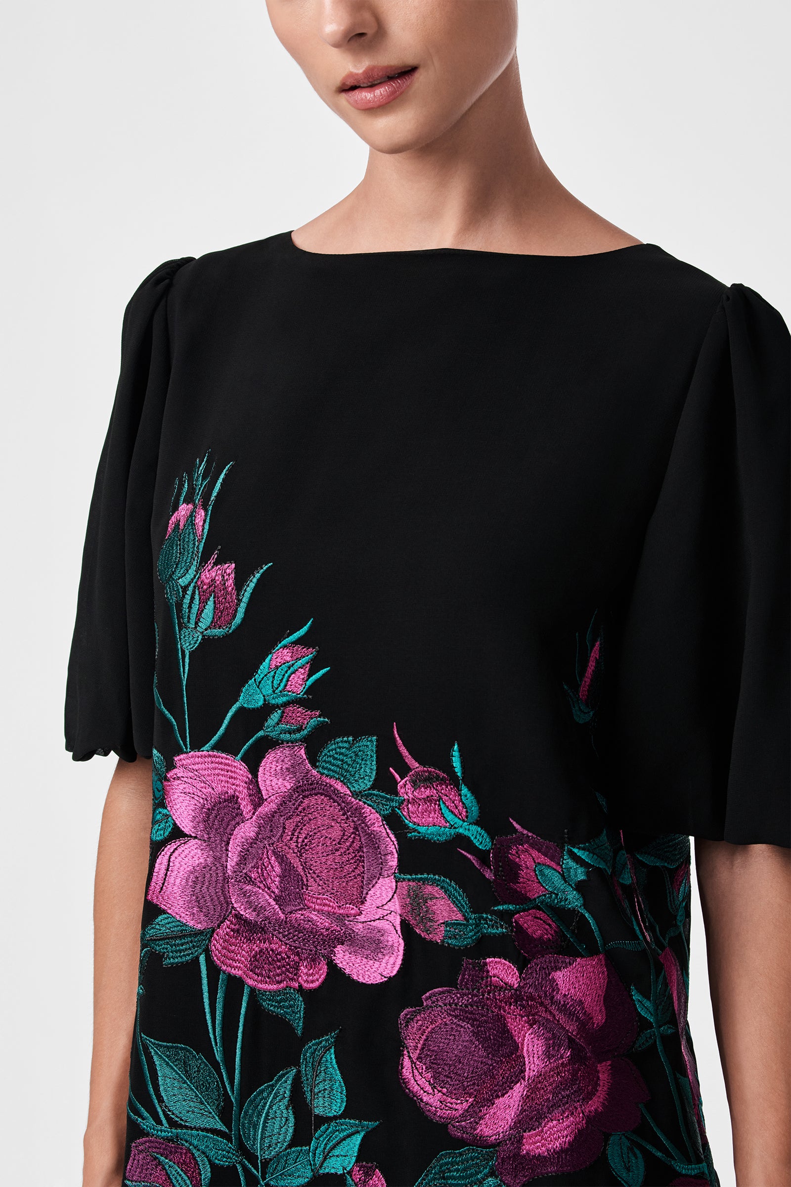 A woman models the Luv Dress—a black, short-sleeved dress featuring bright pink and green floral embroidery on the front. The image shows her from shoulders to waist against a neutral background.