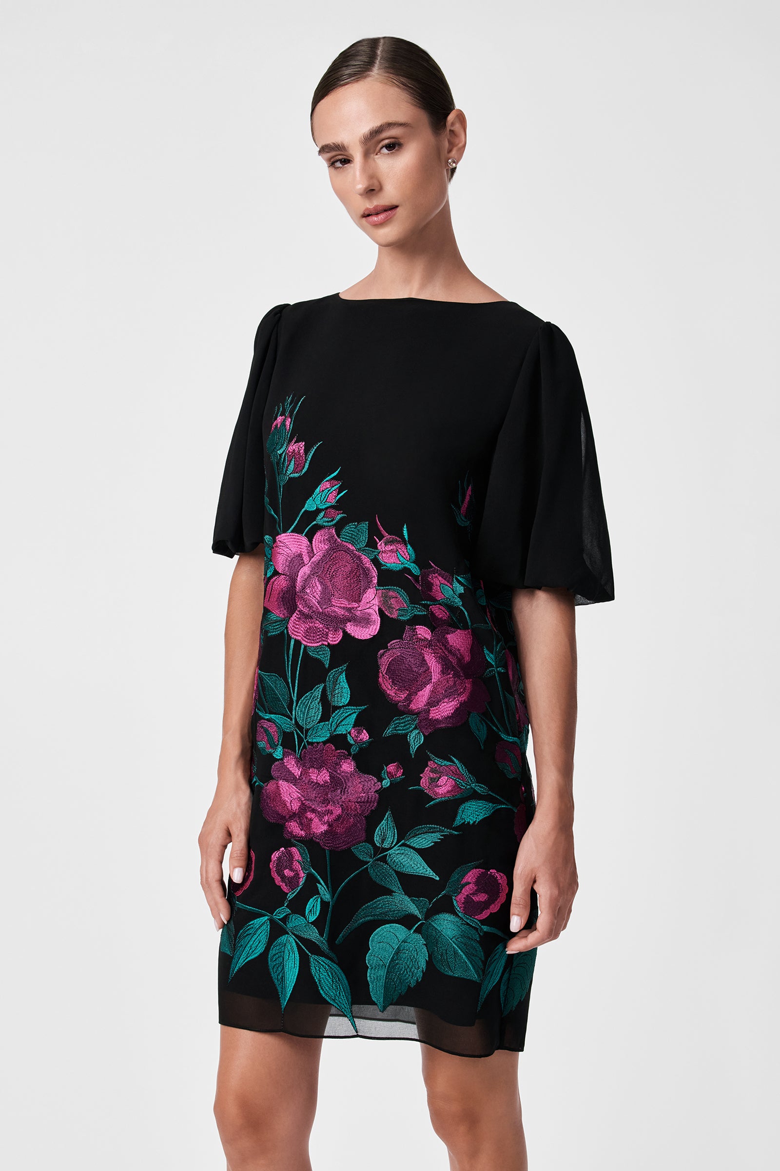 A woman poses against a light gray background wearing the Luv Dress, a black dress with short, flowy sleeves and a vibrant pink floral pattern with green leaves on the front.