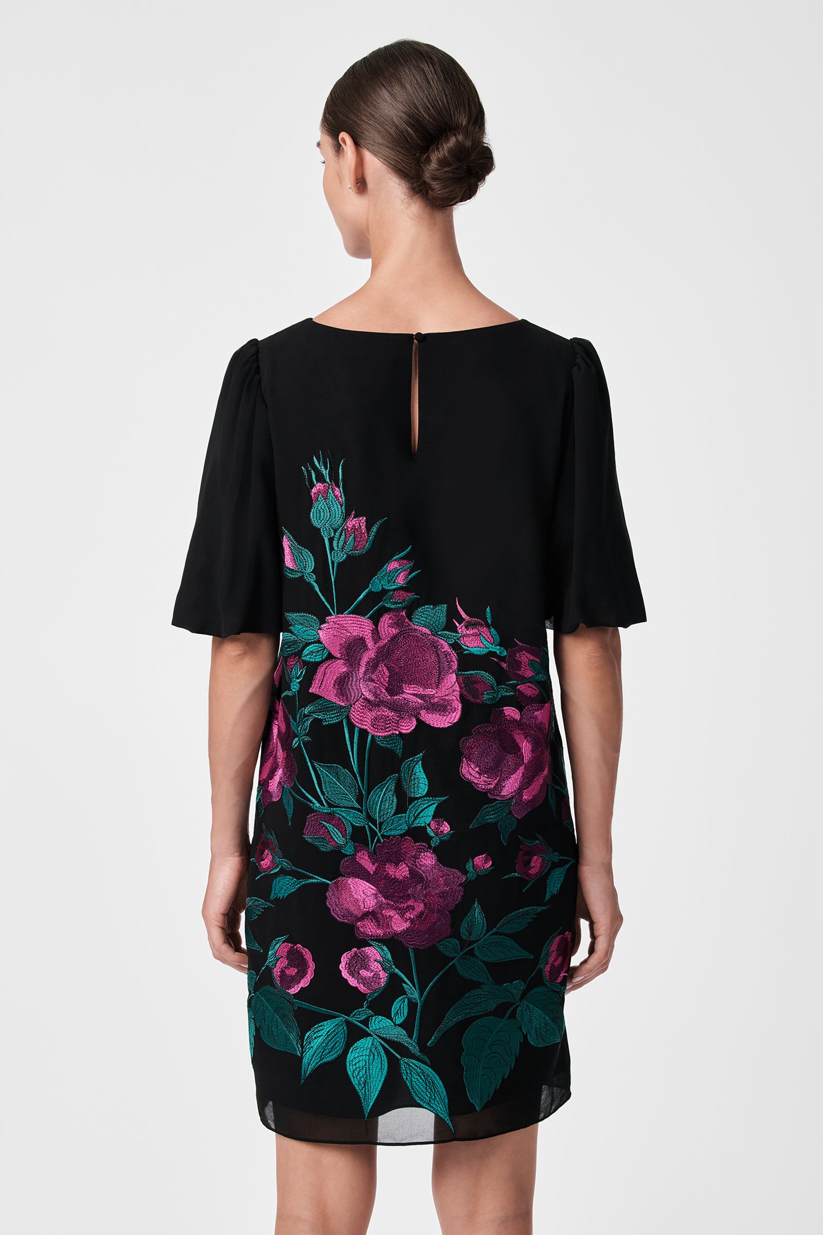 A woman with brown hair in a low bun is seen from the back wearing the Luv Dress—a black dress featuring short sleeves and bold pink and green floral patterns on the lower half, set against a plain light background.