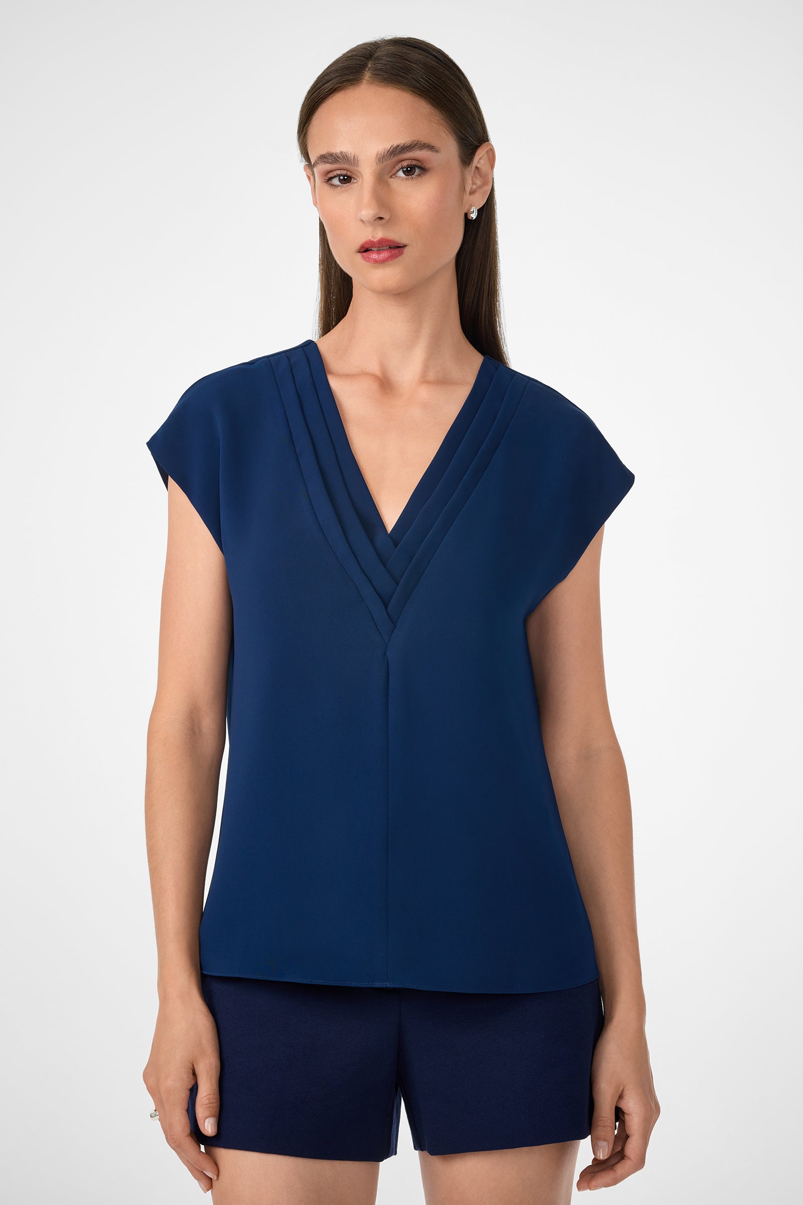 A woman with straight brown hair wears the Zula Top, a navy blue V-neck blouse with short sleeves, paired with matching navy shorts, against a plain light background.