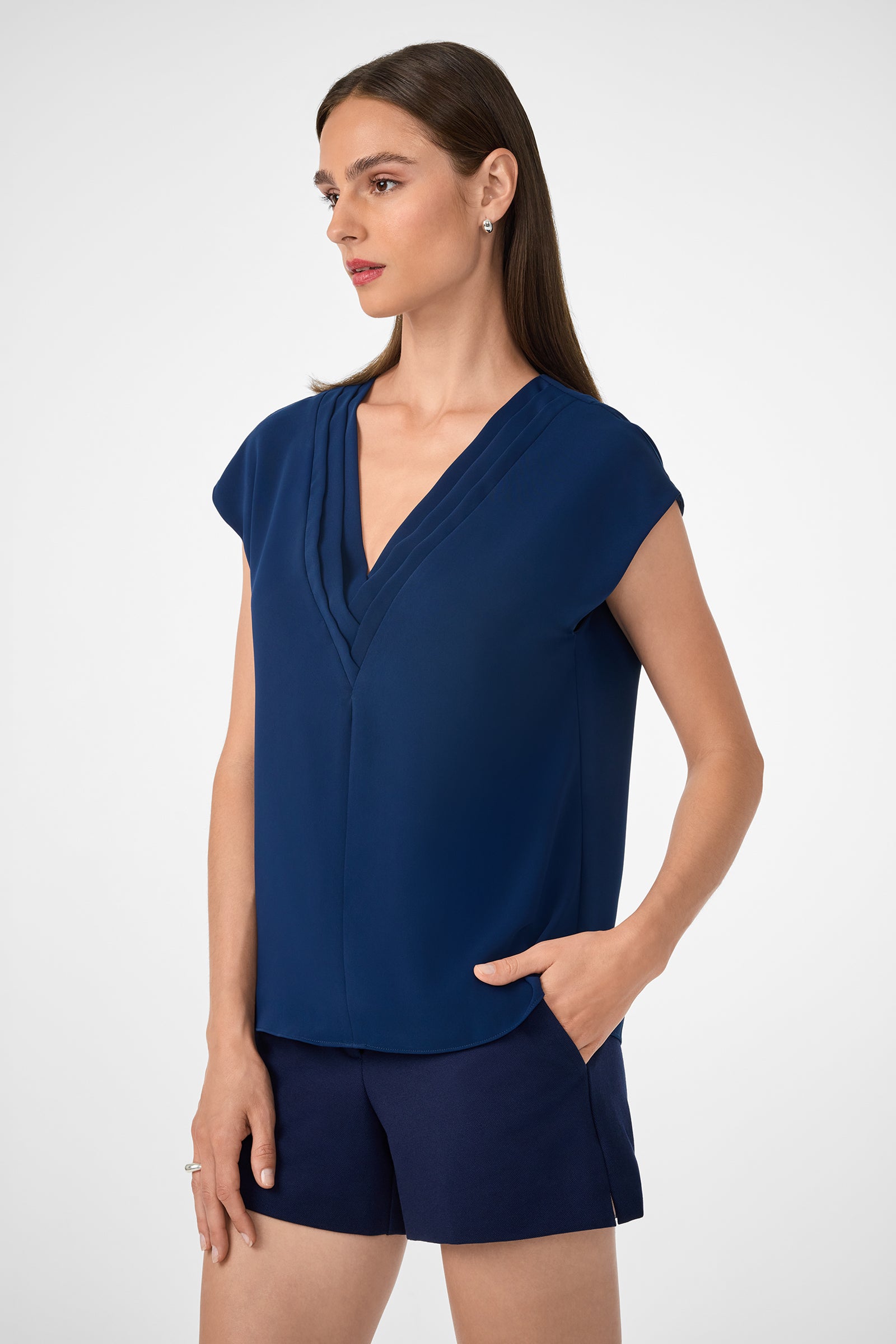 A woman with straight brown hair wears the Zula Top—a navy blue, short-sleeve V-neck blouse—and dark navy shorts. She stands with one hand in her pocket against a plain light background, looking to the side.