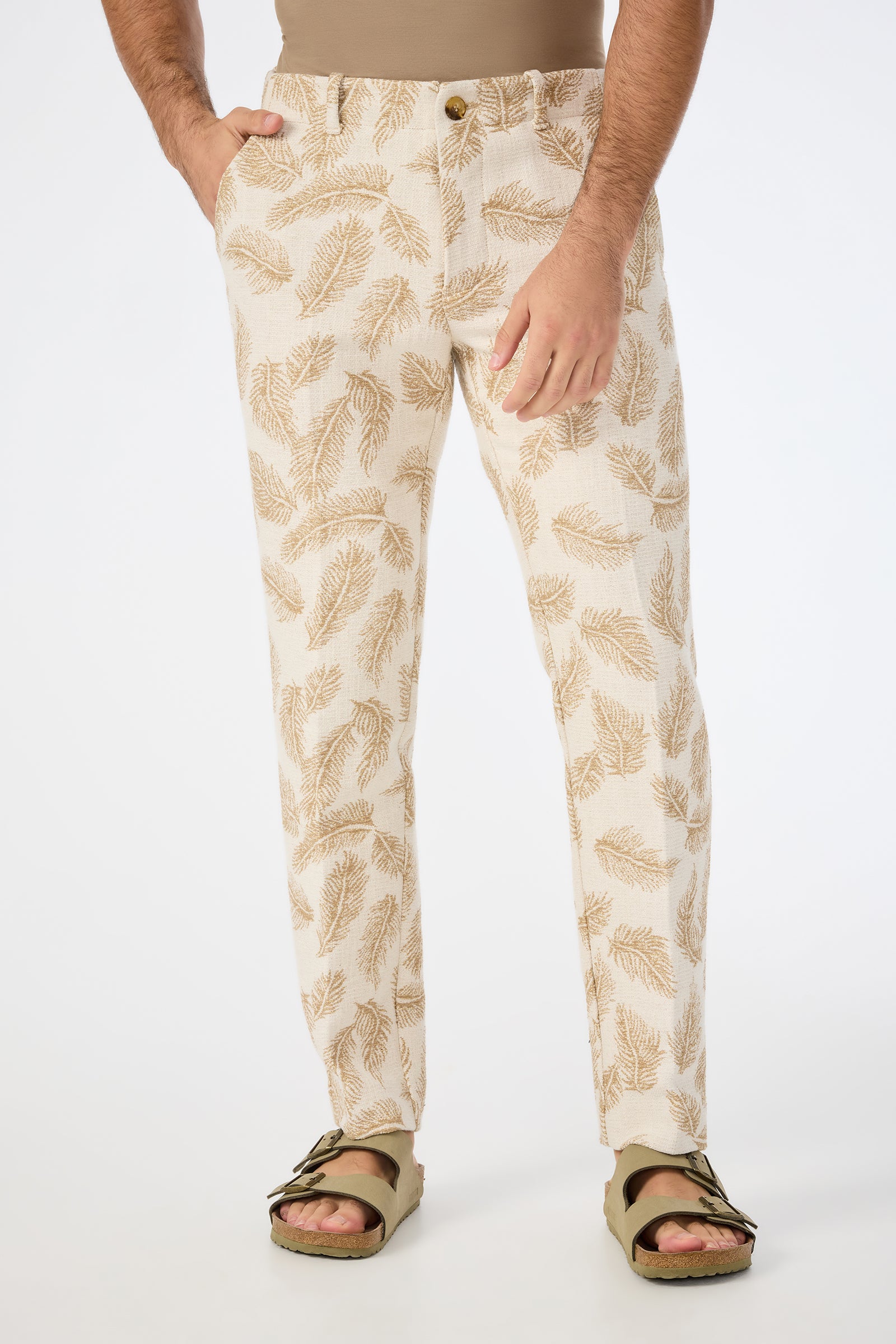 Wearing the Alex Slim Trouser in Desert Fronds Jacquard, a beige shirt tucked in, and olive green sandals, a person stands against a plain white background with hands in pockets.