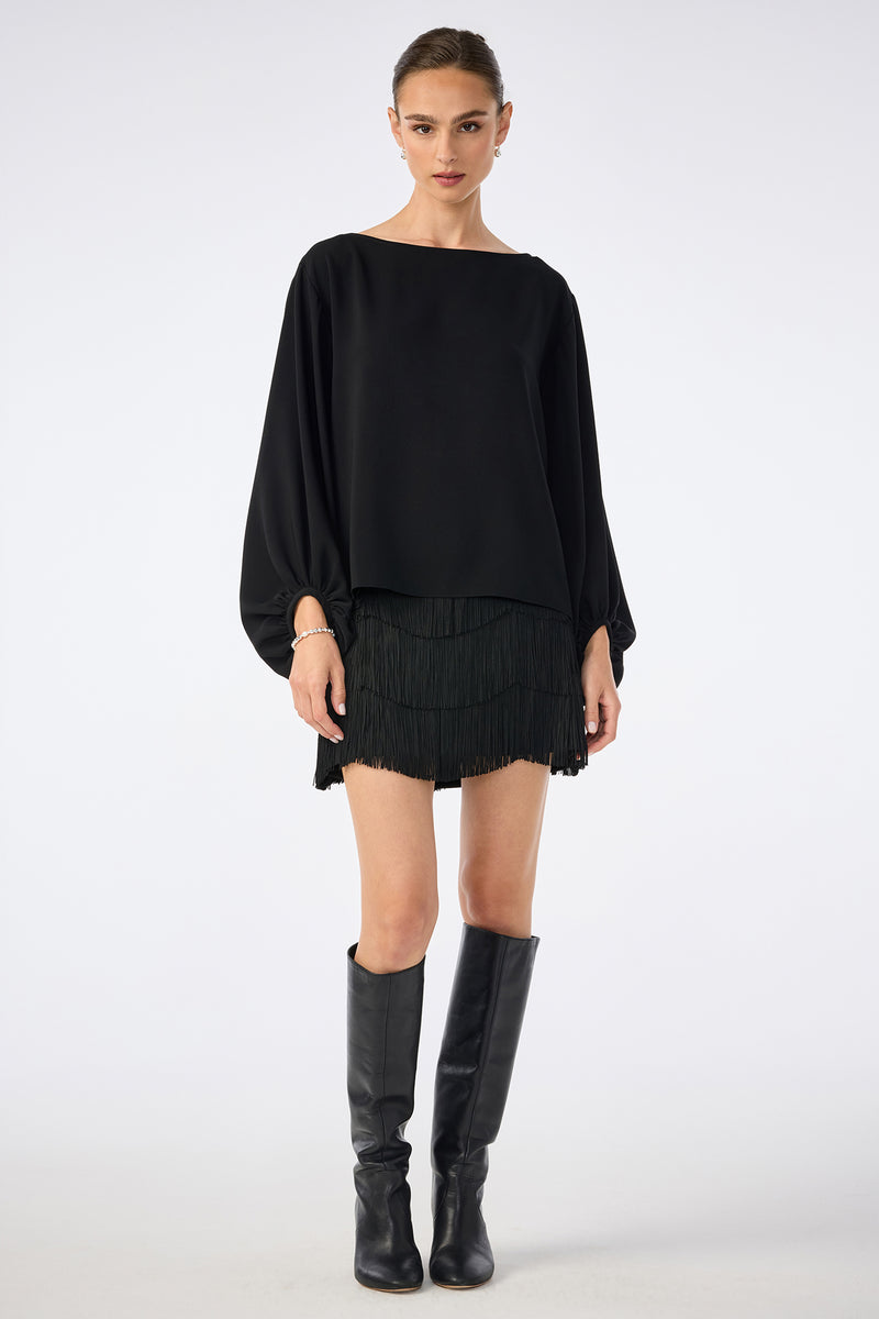A woman wearing a loose black long-sleeve top, the Chocolat Skirt—a black fringed mini skirt—and knee-high black boots stands against a plain light background.