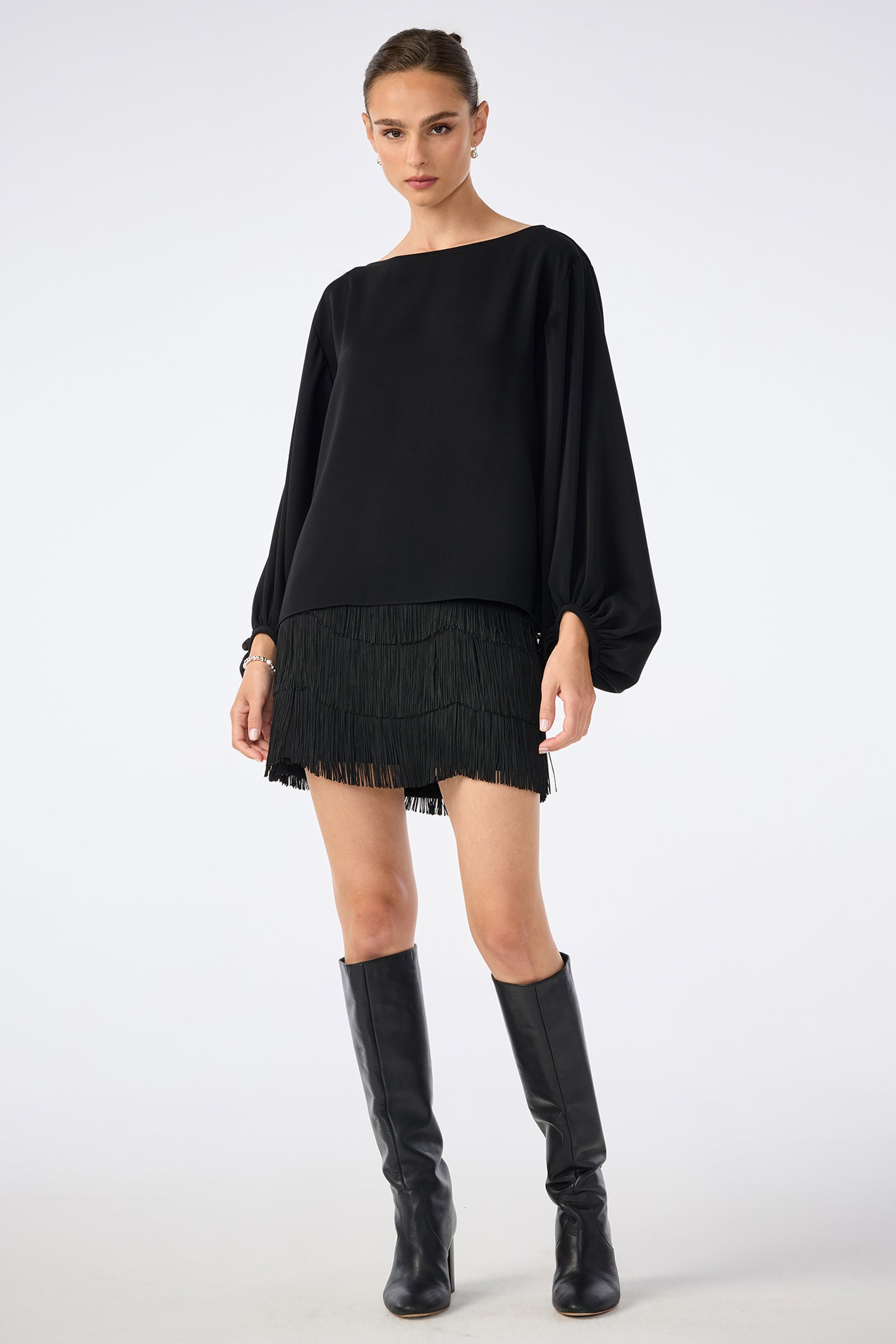 A woman stands against a plain background wearing the URSA 2 TOP, a loose black long-sleeved shirt, paired with a black fringe mini skirt made from 100% polyester and black knee-high boots. She looks straight ahead with a neutral expression.