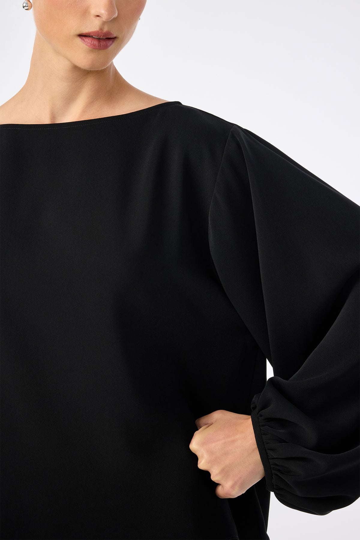 A woman models the URSA 2 TOP, a loose-fitting black long-sleeve blouse with puffed sleeves made from 100% polyester, shown from shoulders to waist, one hand on her hip, against a plain light background.