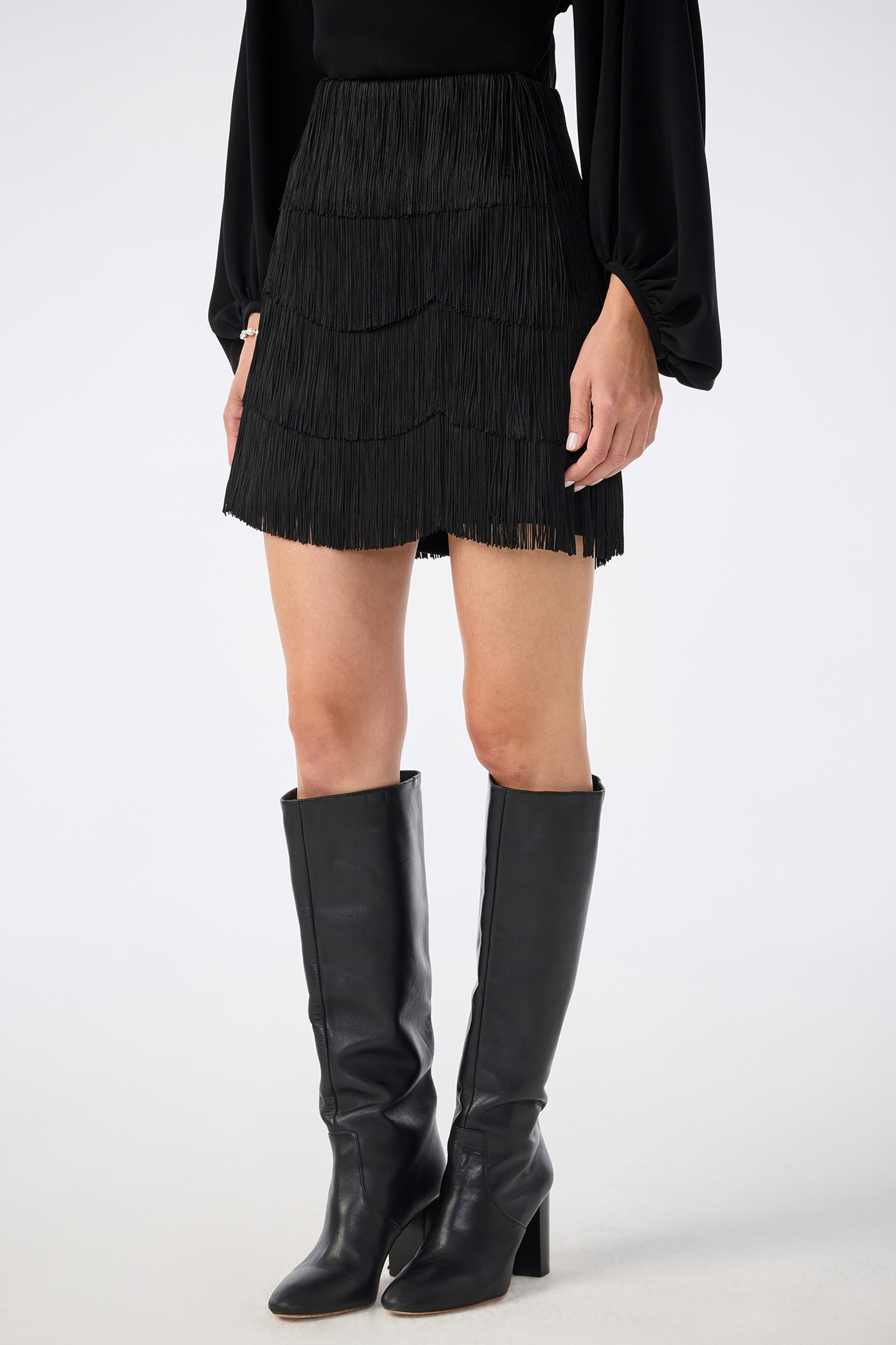 A person wearing a black long-sleeve top, the Chocolat Skirt (a black tiered fringe mini skirt), and knee-high black heeled boots, standing against a plain light background.