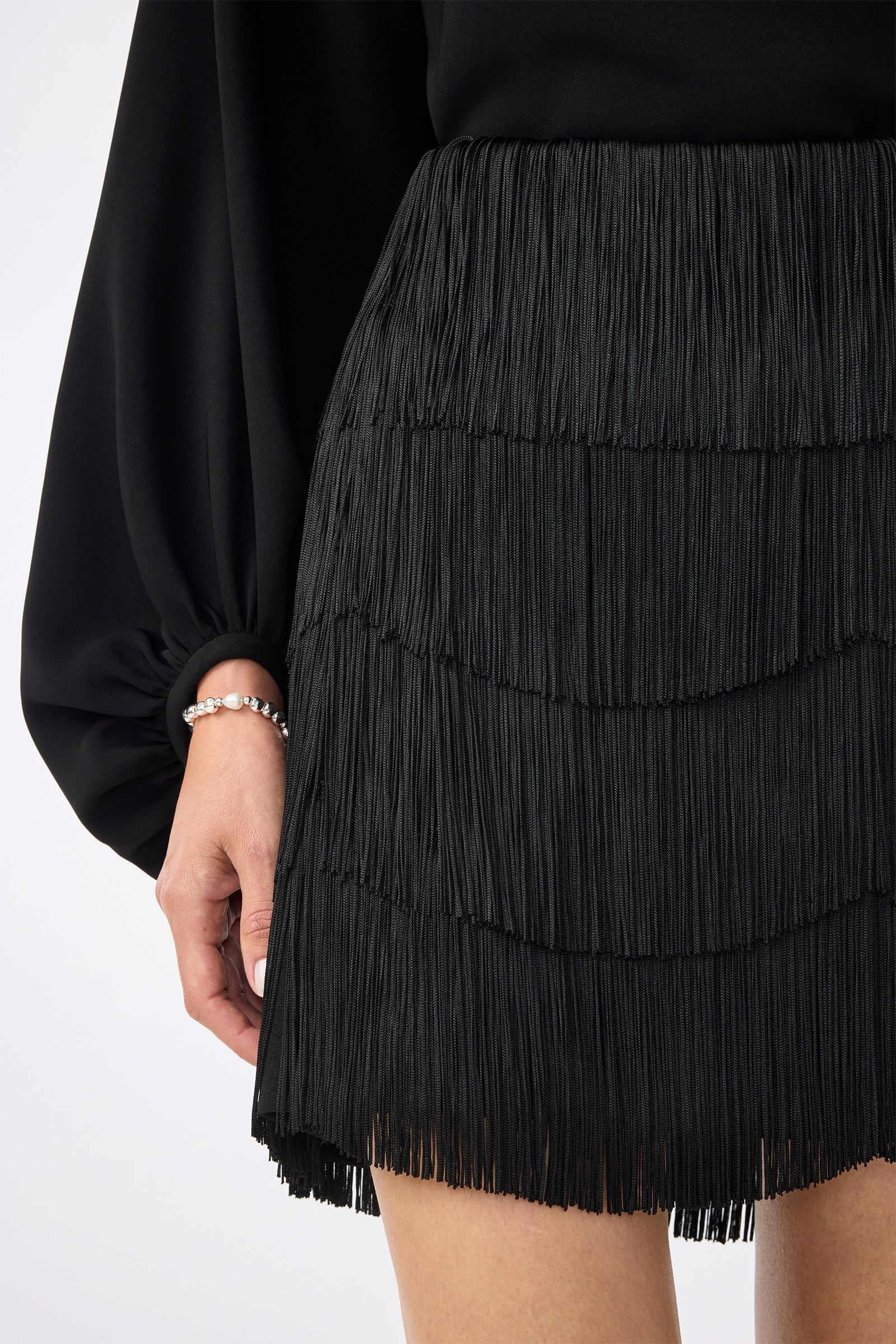 A person models the Chocolat Skirt—a black, fringed design—paired with a black long-sleeve top. They wear a silver bracelet on their left hand, standing against a plain, light background.