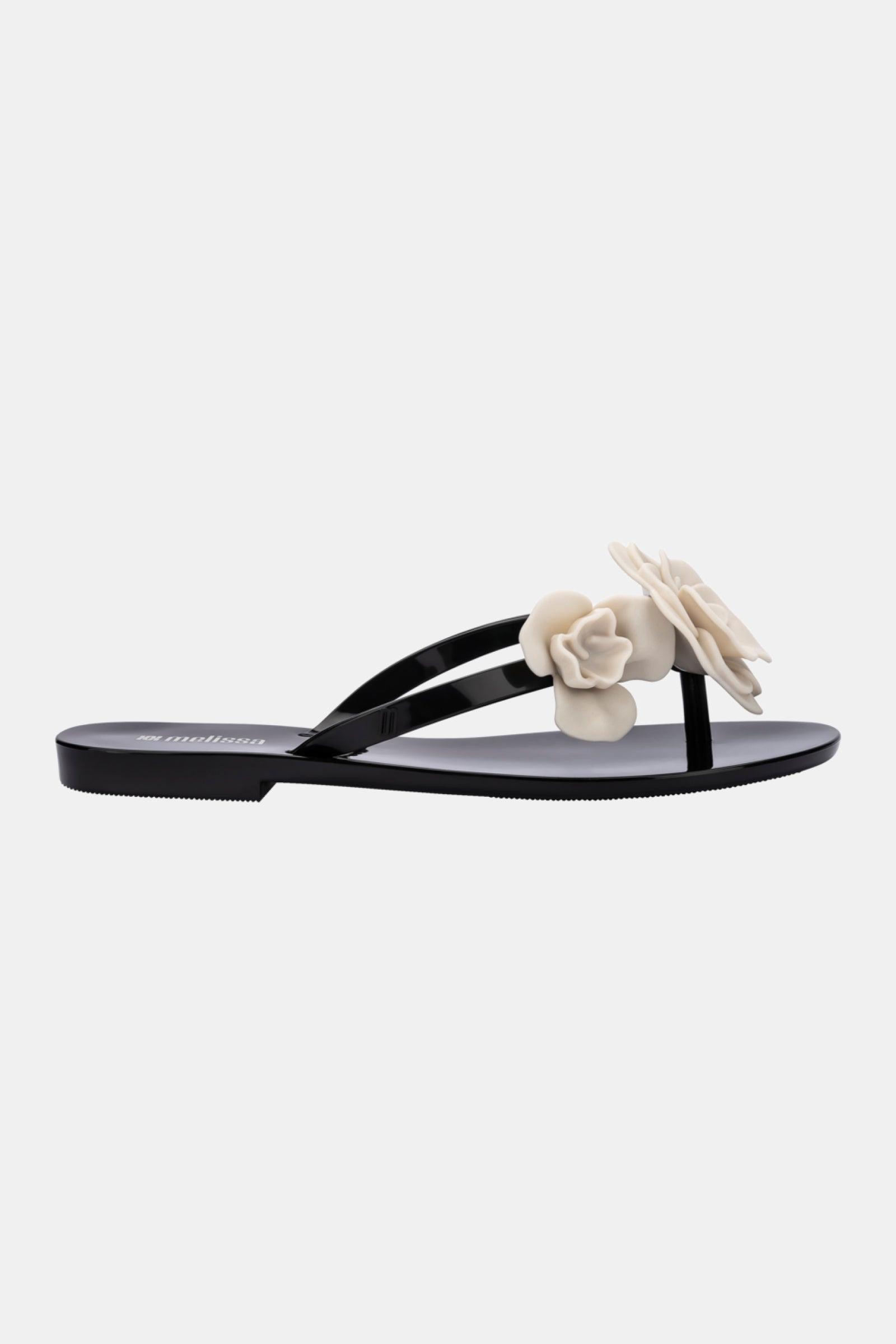 The Melissa Harmonic Petals Sandal is a black flat sandal with thin straps and two beige flower embellishments on top, shown against a plain white background.