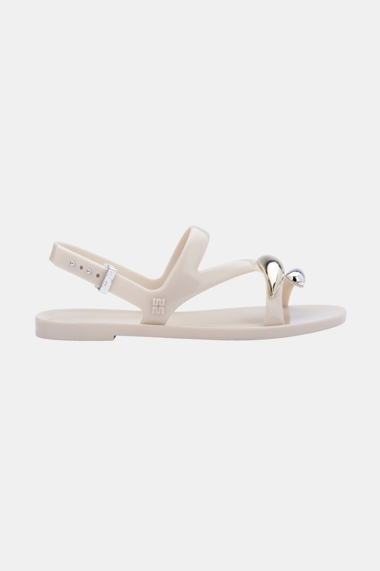 The Melissa Iris Sandal is a beige flat with an adjustable ankle strap and a metallic silver accent over the toe loop, shown against a plain white background.