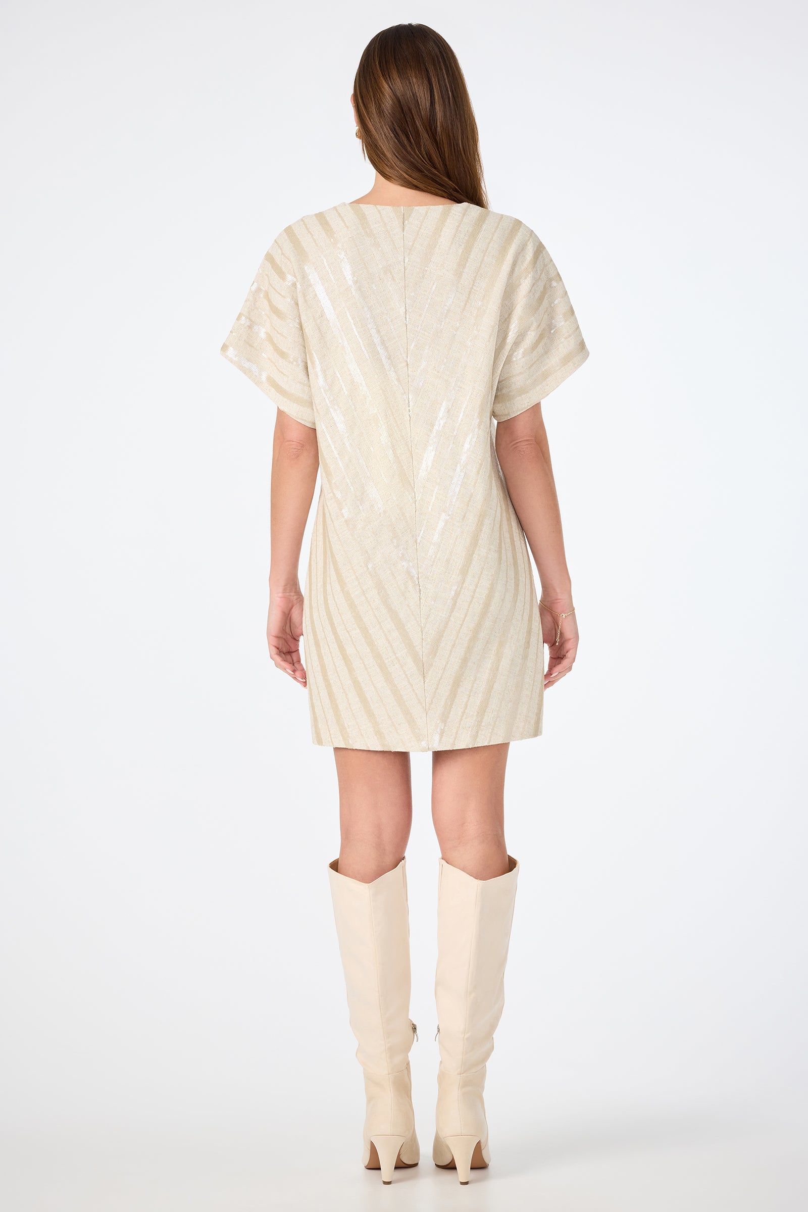 A woman with long brown hair stands with her back to the camera against a white background, wearing the Elisabetta Short-Sleeve Dress in cream and knee-high beige boots.