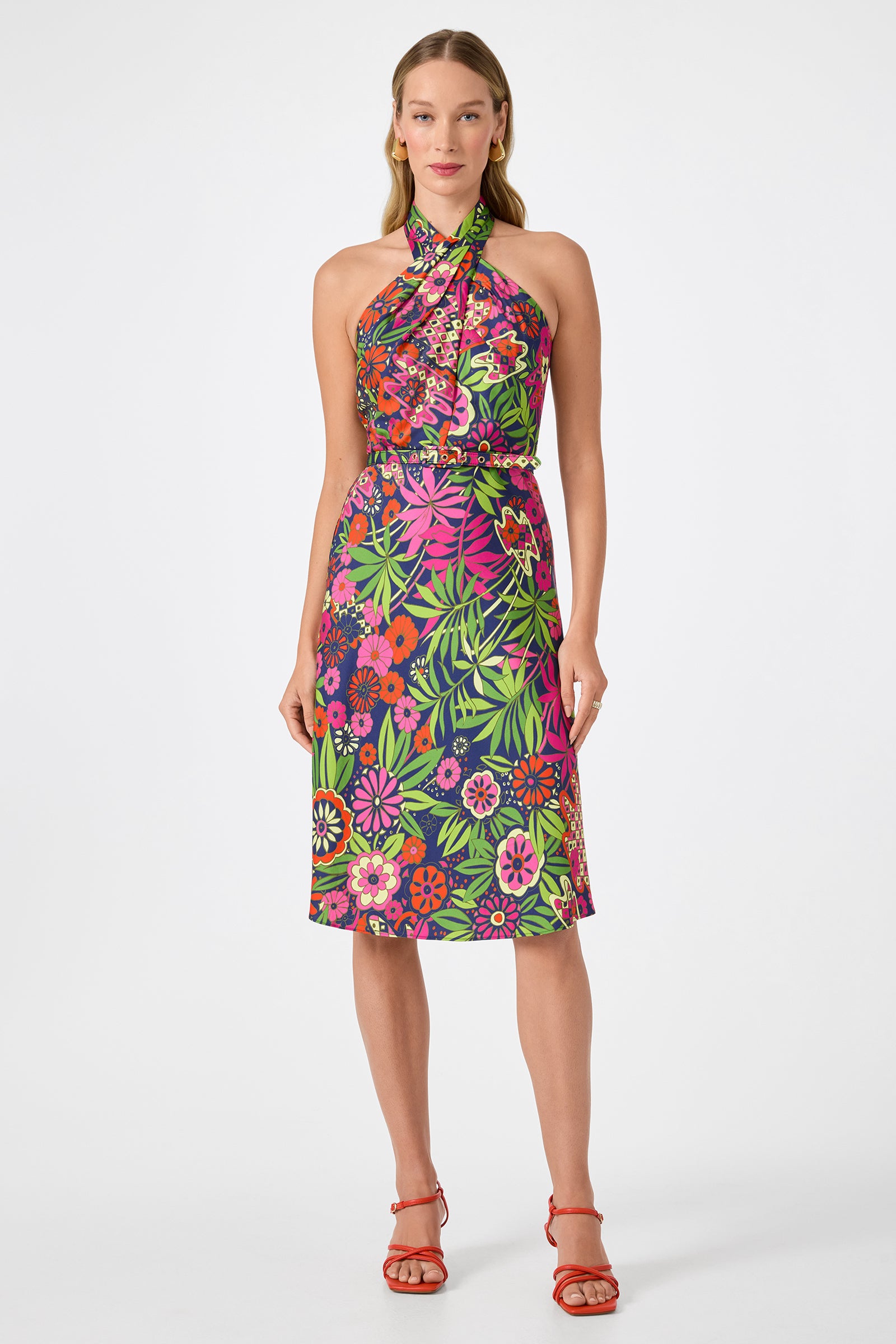 A woman models the Muri Cross-Halter Midi Dress in Fumi Floral, featuring a vibrant pink, green, and purple pattern. She wears red strappy sandals and stands against a plain white background.