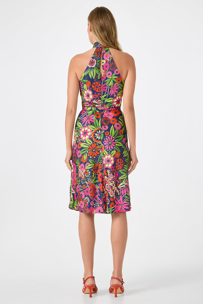 A woman stands facing away, wearing the Muri Cross-Halter Midi Dress - Fumi Floral, a colorful floral halter-neck dress thats fitted at the waist and knee-length, paired with red heeled sandals. The background is plain white.