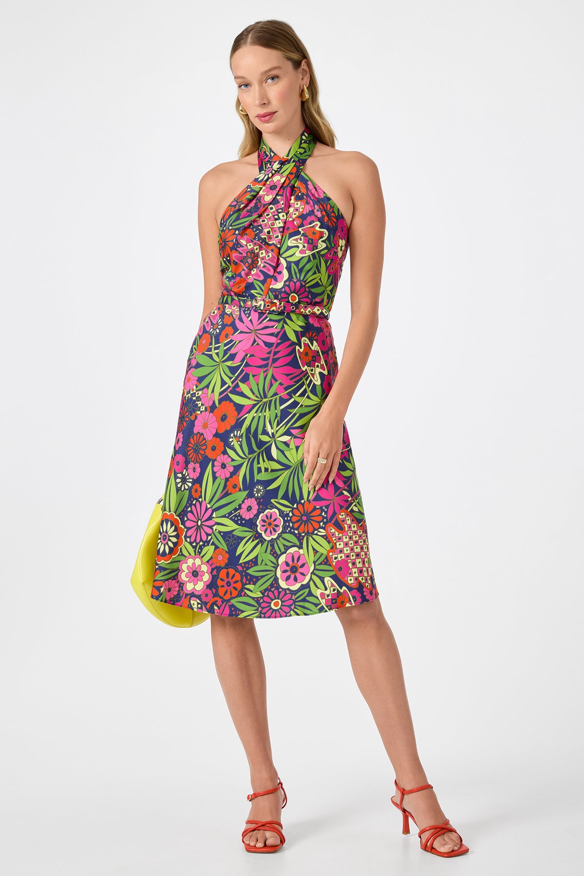 Woman wearing the Muri Cross-Halter Midi Dress in Fumi Floral, featuring a vibrant red, pink, green, and yellow floral pattern with a cross-halter neckline, paired with red heeled sandals against a plain white background.