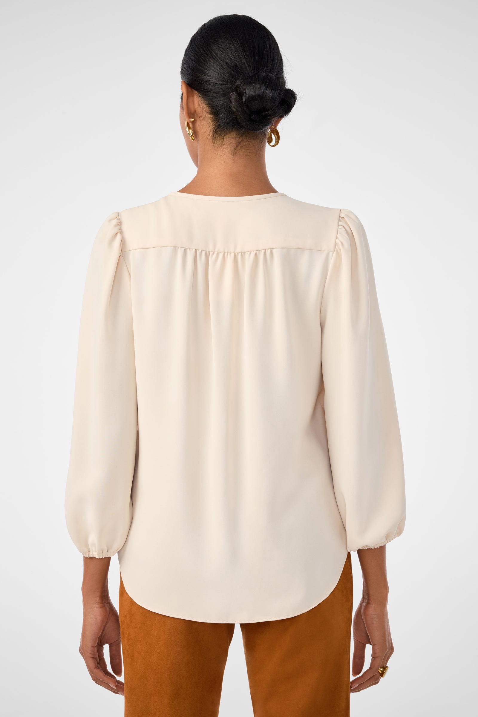A woman with dark hair in a low bun, wearing gold hoop earrings and the cream-colored Morabi Split-Neck Top, stands facing away from the camera against a plain background.