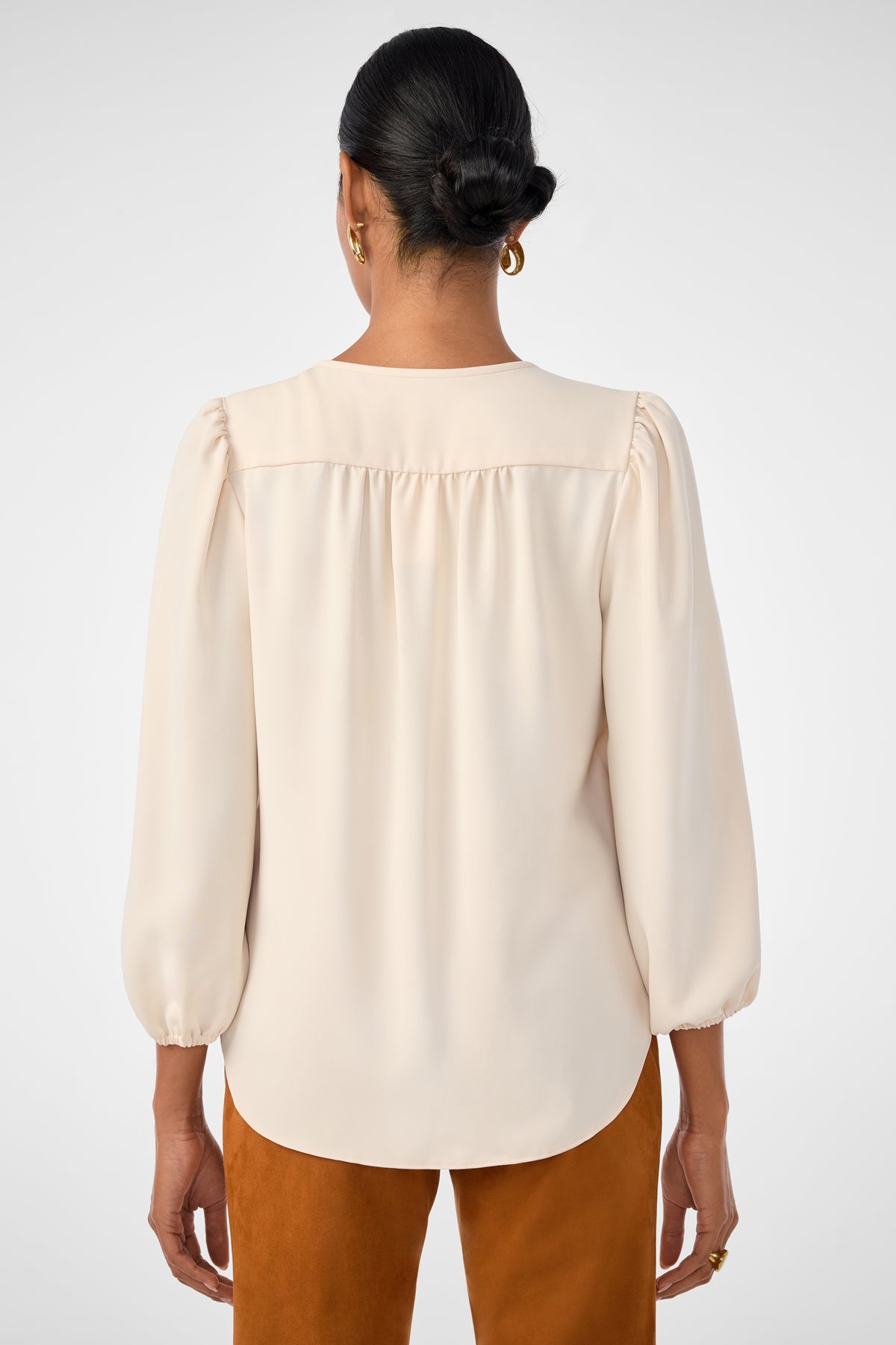 A woman with dark hair in a low bun, wearing gold hoop earrings and the cream-colored Morabi Split-Neck Top, stands facing away from the camera against a plain background.