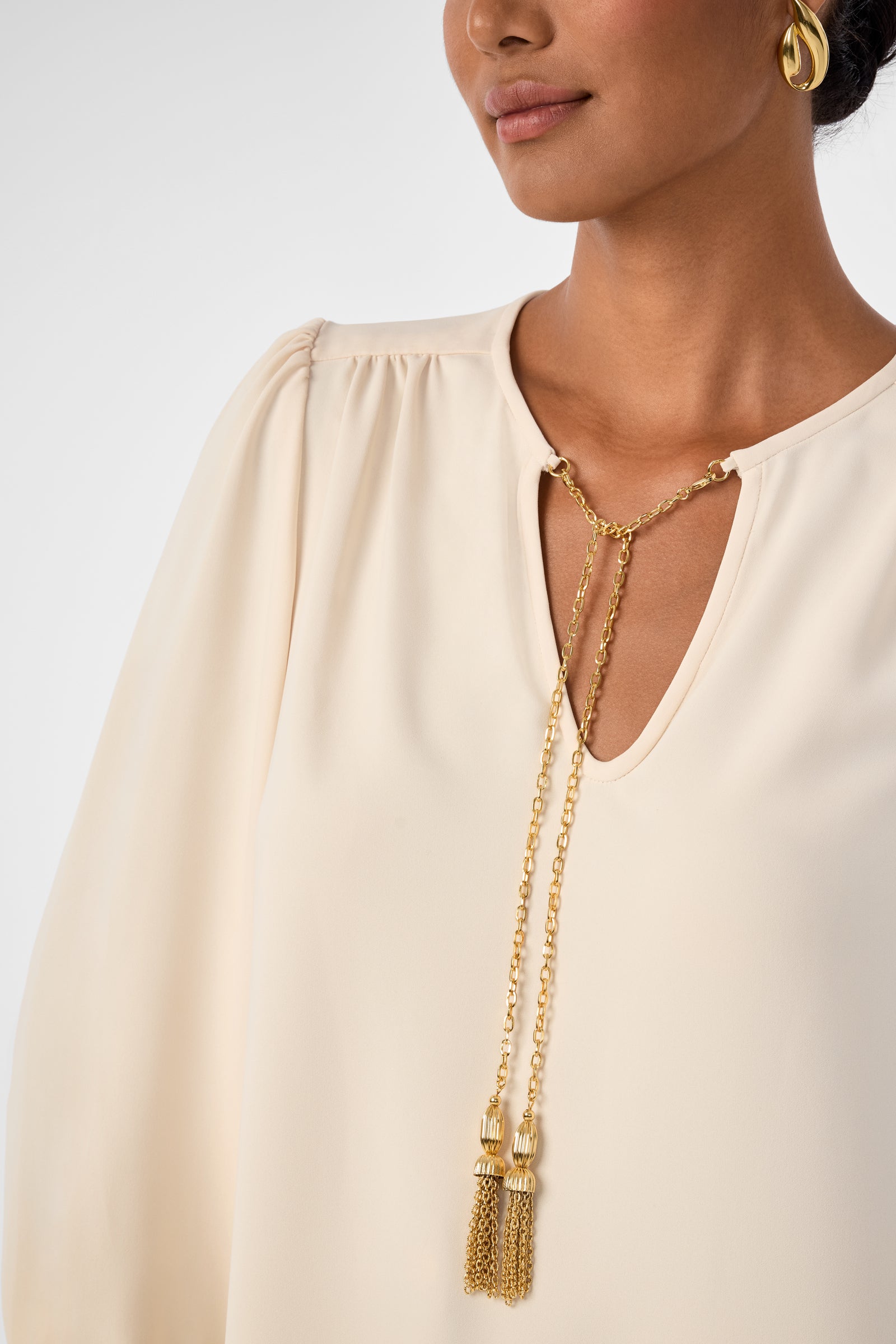 A woman wearing the Morabi Split-Neck Top in cream, accessorized with a gold chain tassel necklace and matching earrings. Only the lower half of her face is visible.