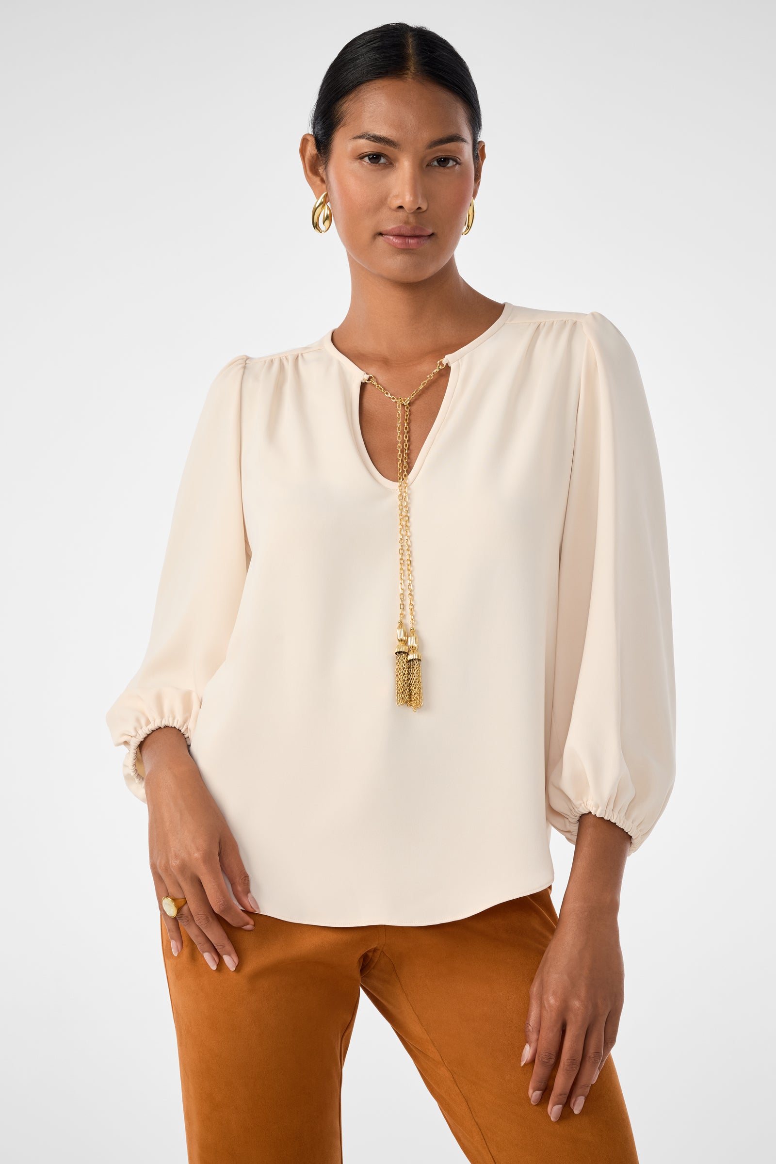 A woman wearing the Morabi Split-Neck Top in Poplar with long sleeves and a split neckline, styled with gold jewelry and ochre pants, stands against a plain light background.