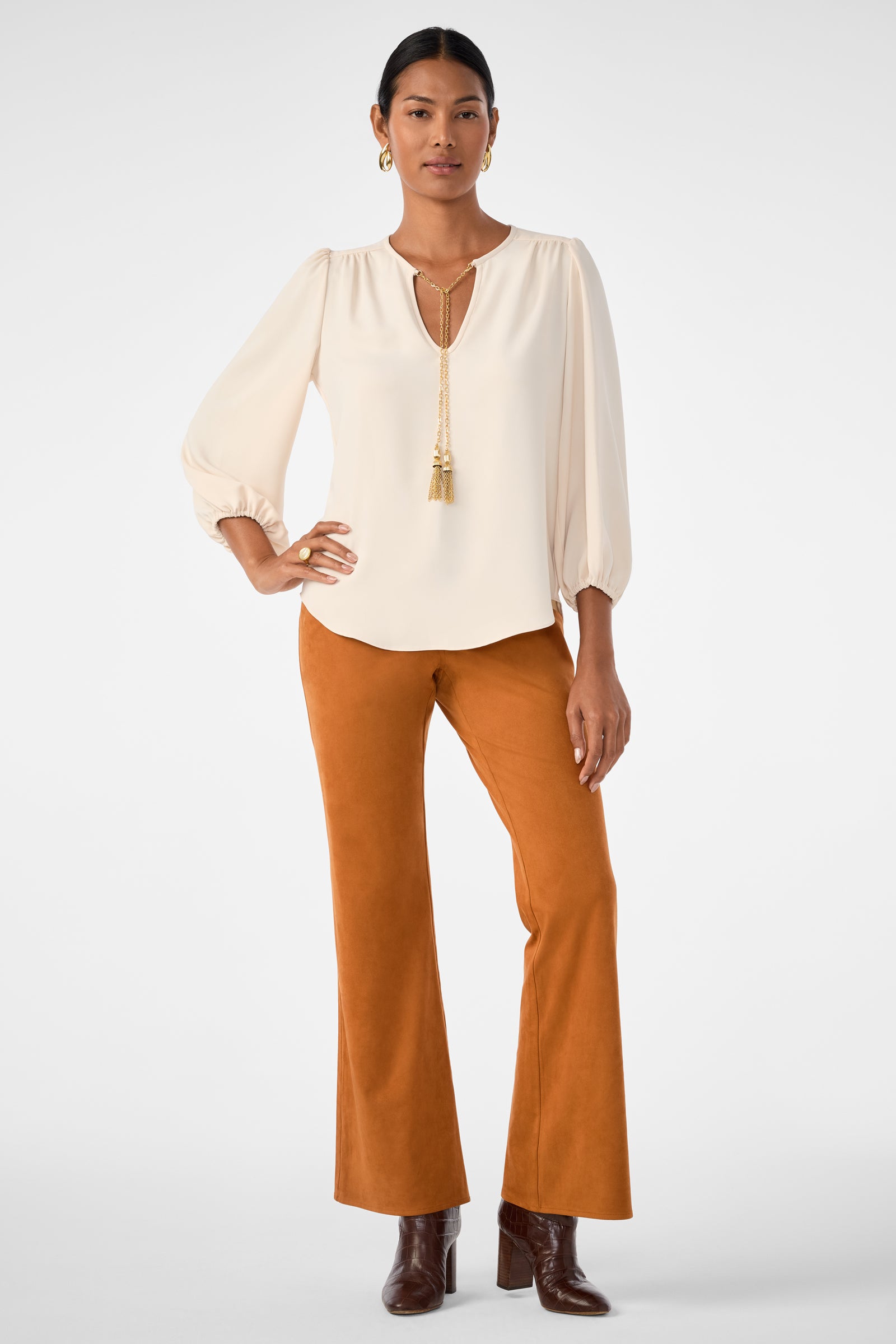 A woman stands confidently in the Morabi Split-Neck Top with three-quarter sleeves, rust flared pants, brown heeled boots, gold hoops, and a long tassel necklace against a plain background.