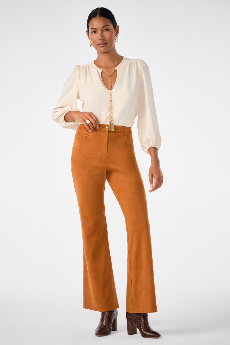 A woman wears the Apple Bottom Pant, a high-waisted rust-colored flared style, paired with a cream long-sleeve blouse, brown heeled boots, and gold jewelry against a plain white background.