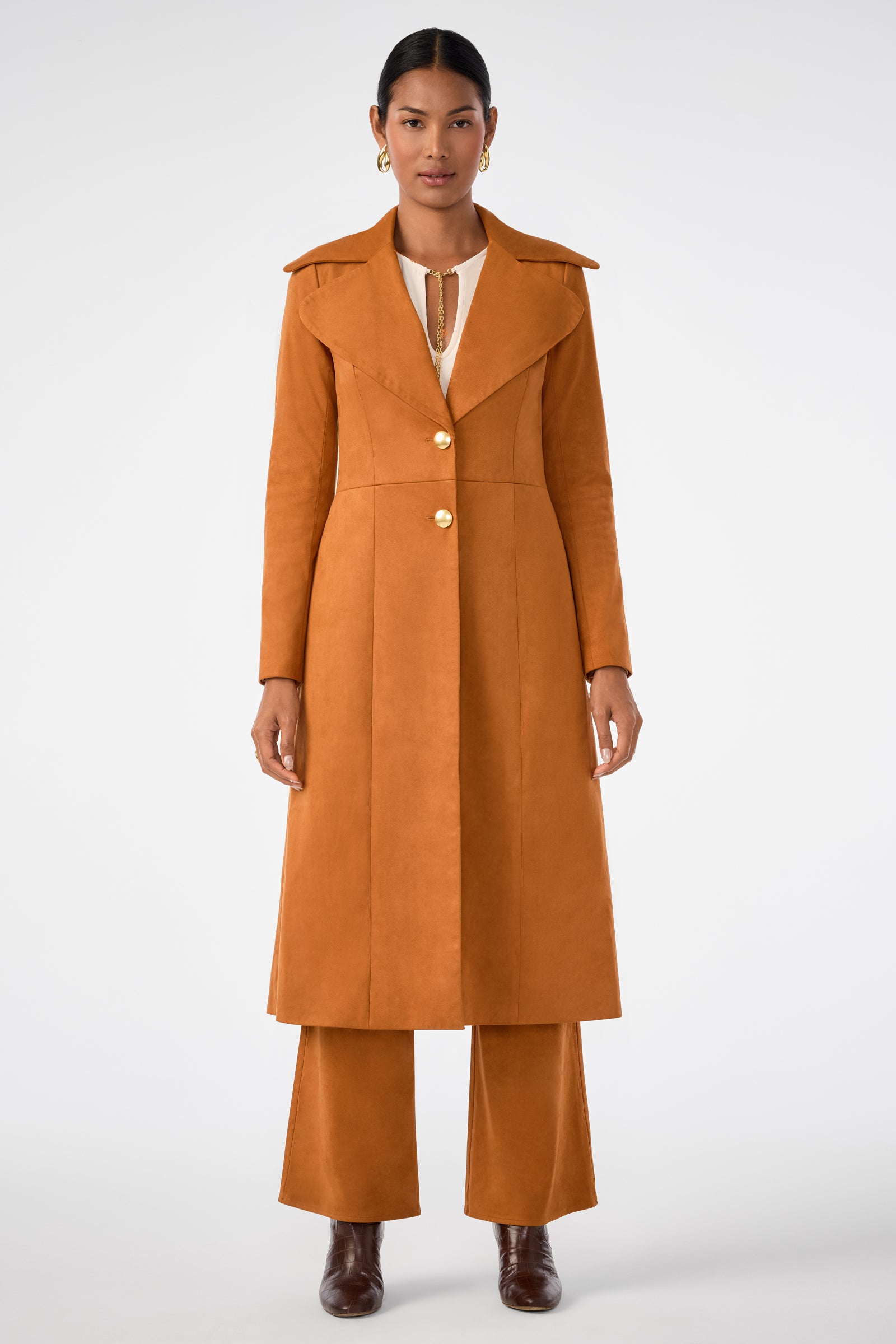 A woman faces forward in the Starflower Coat, a long rust-orange piece with gold buttons, styled over matching wide-leg pants and ankle boots. She wears gold hoop earrings, has her hair pulled back, and stands against a plain white background.