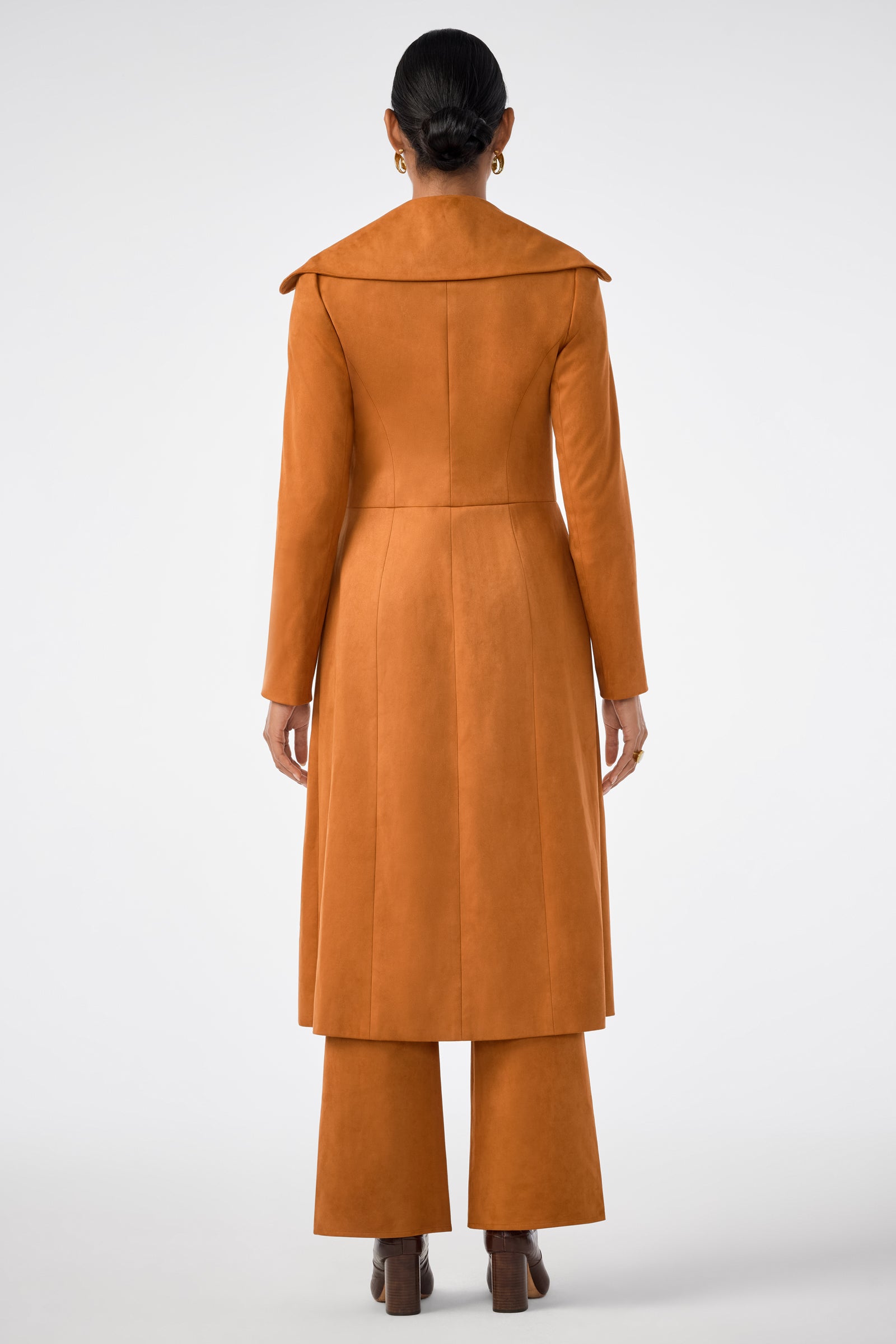 A person stands facing away, wearing the Starflower Coat—a long, burnt orange piece with a wide collar—paired with matching wide-leg pants and heeled shoes against a plain light background.