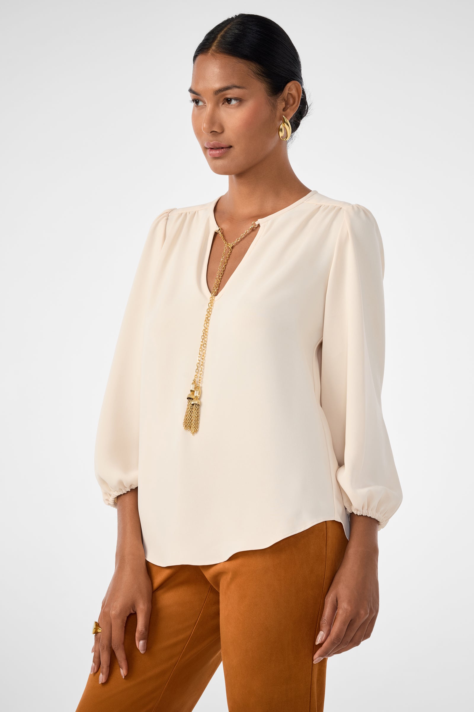 A woman wears the Morabi Split-Neck Top, a cream-colored long-sleeve blouse with a V-neck, styled with rust pants. Her dark hair is pulled back and she poses against a plain light background, accessorized with a gold tassel necklace.