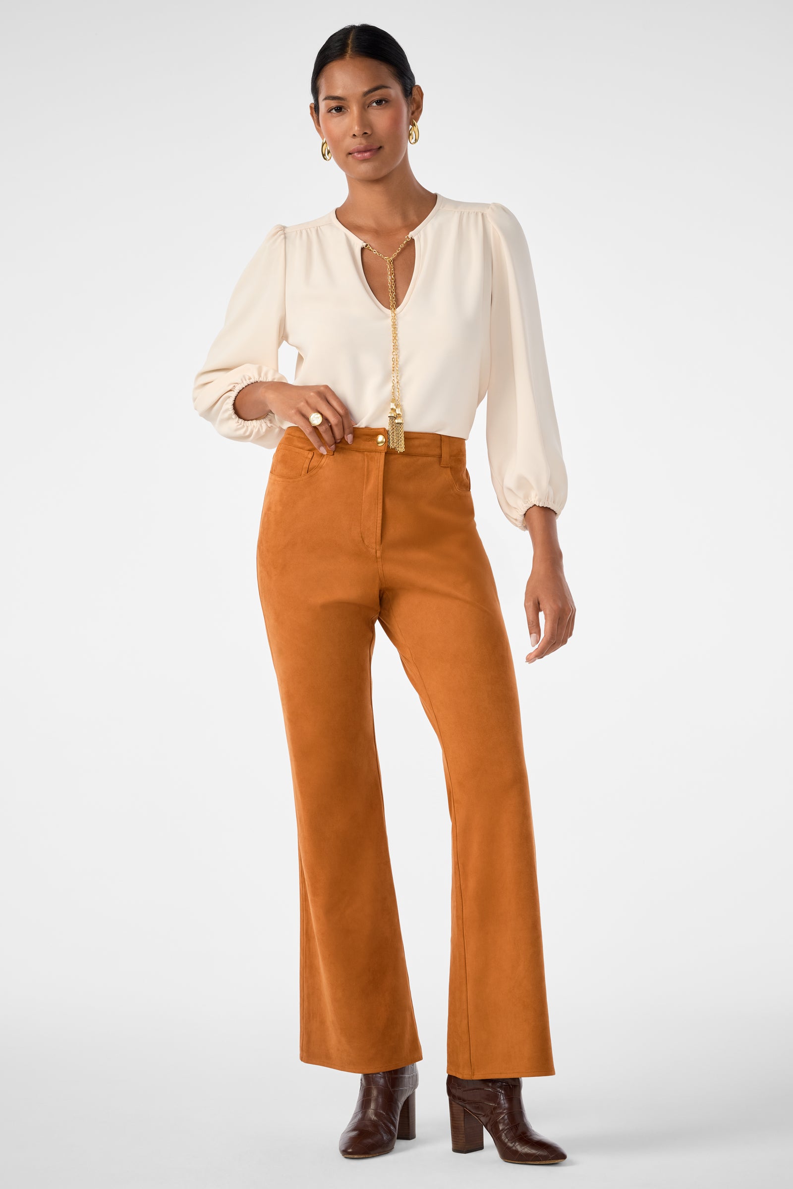 A woman stands against a white background wearing the Morabi Split-Neck Top, rust-colored high-waisted pants, brown heeled boots, and gold hoop earrings. She has one hand on her hip and looks at the camera.