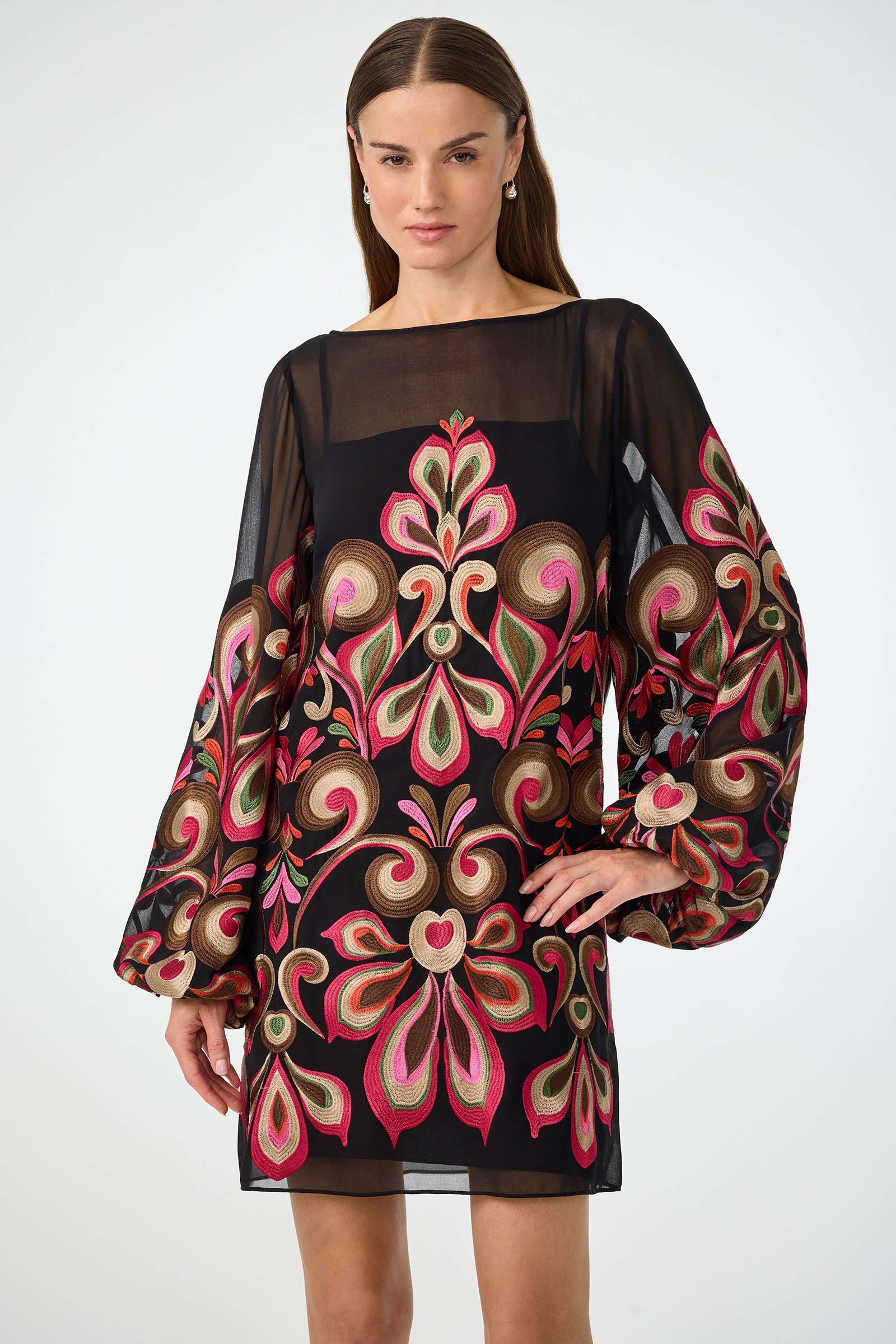 A woman stands with one hand on her hip in the Salim Embroidered Mini Dress—a black, sheer long-sleeve dress featuring bold floral and paisley embroidery in pink, green, gold, and red—against a plain background.