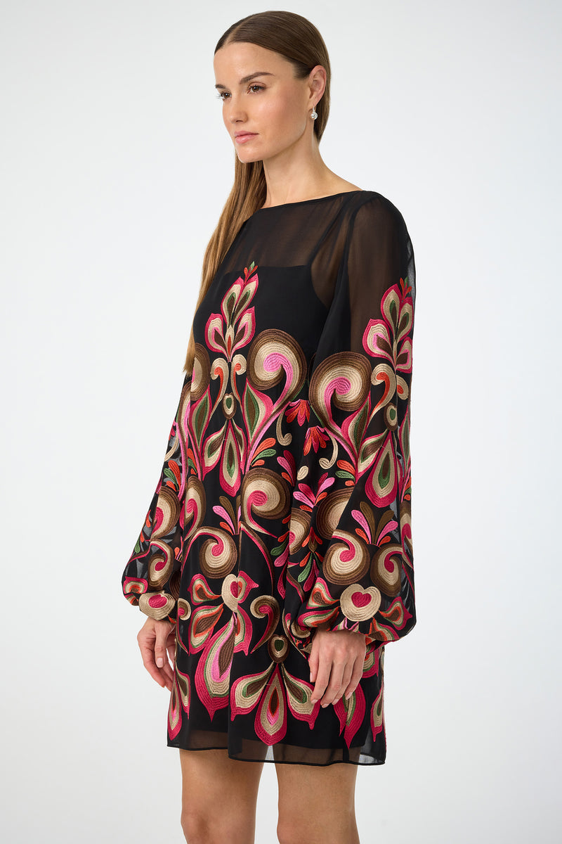 A woman with straight brown hair and a neutral expression models the Salim Embroidered Mini Dress, featuring sheer black fabric, bold colorful floral embroidery, and long billowy sleeves, against a plain white background.
